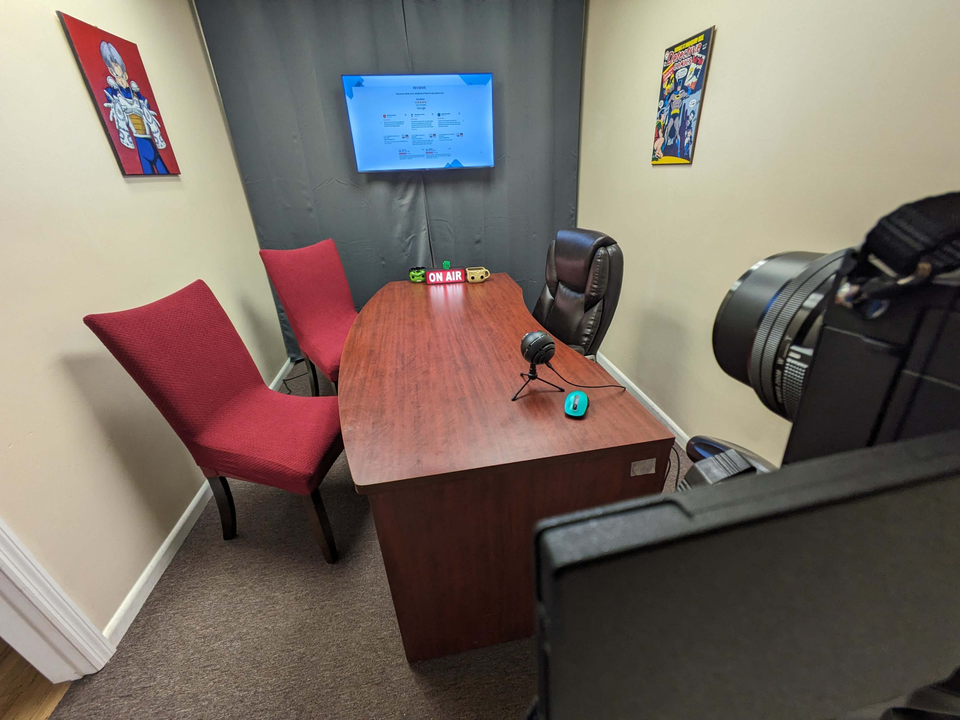 A small recording studio features a wooden table with two red chairs, a microphone, a green indicator light, and a wall-mounted screen displaying information, all set against a neutral-colored background.