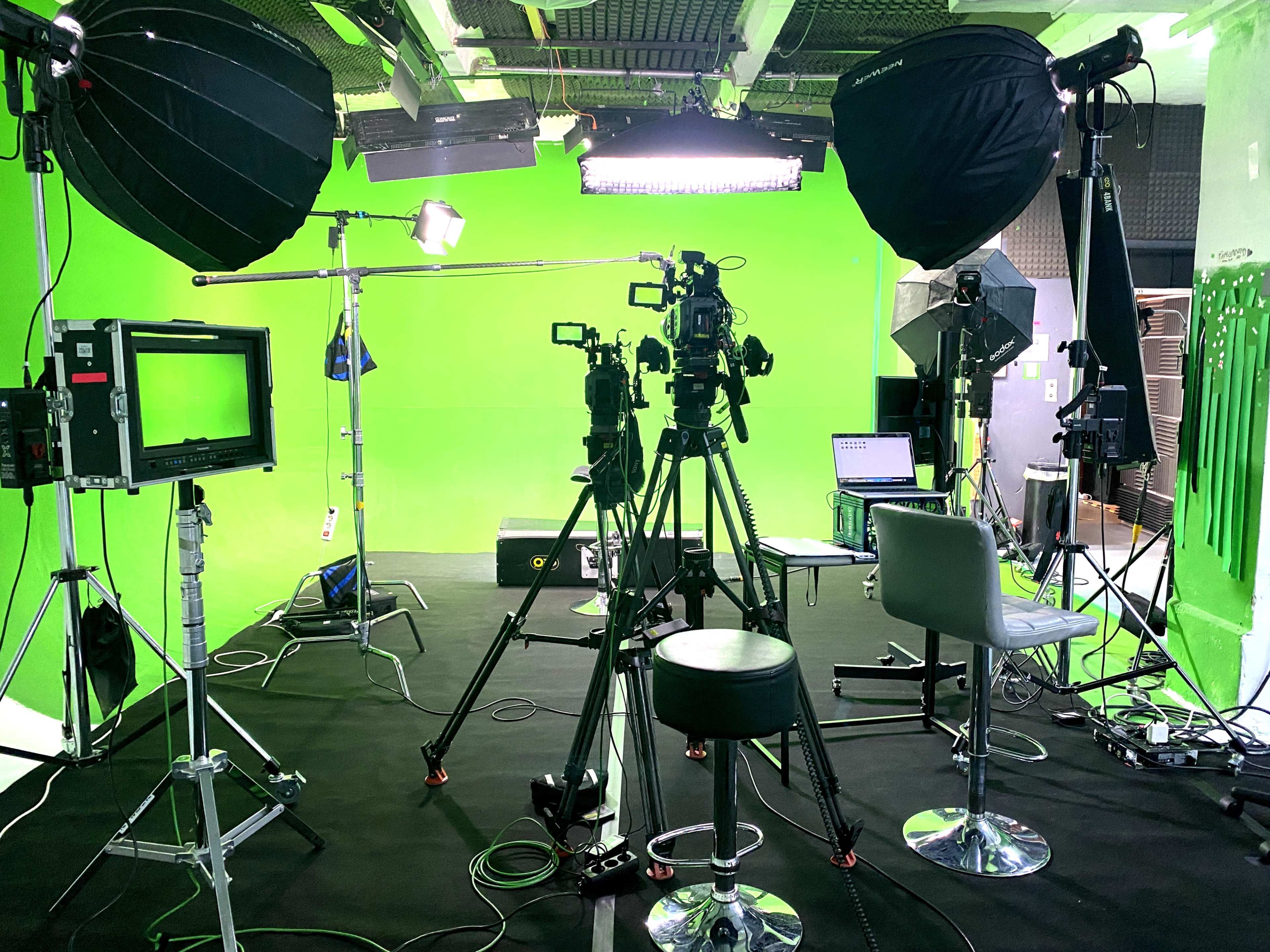 The image shows a television studio setup featuring multiple cameras, lighting equipment, and a green screen backdrop.