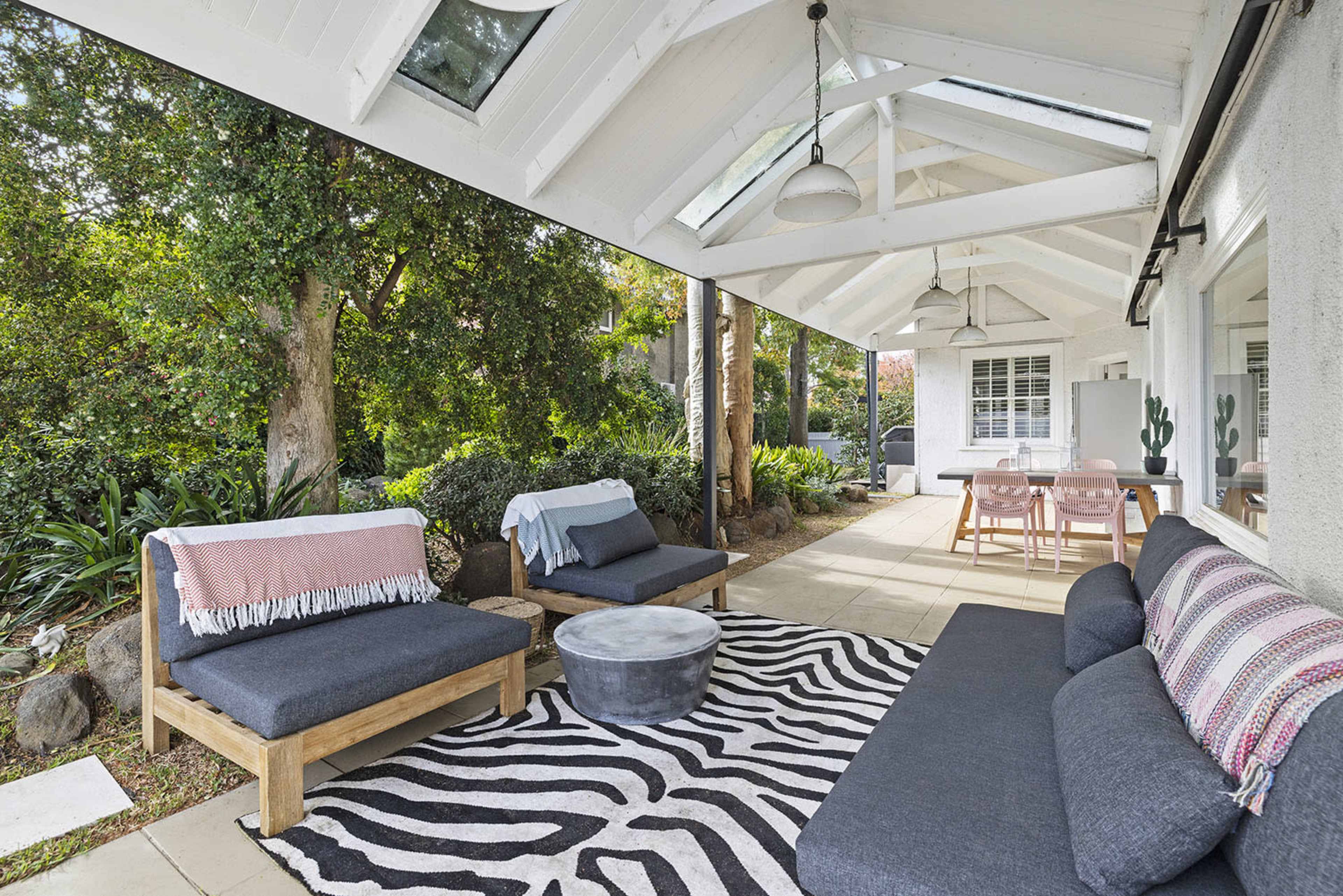 A modern outdoor living space features a seating area with wooden couches, a round coffee table, and a striped rug under a vaulted ceiling.
