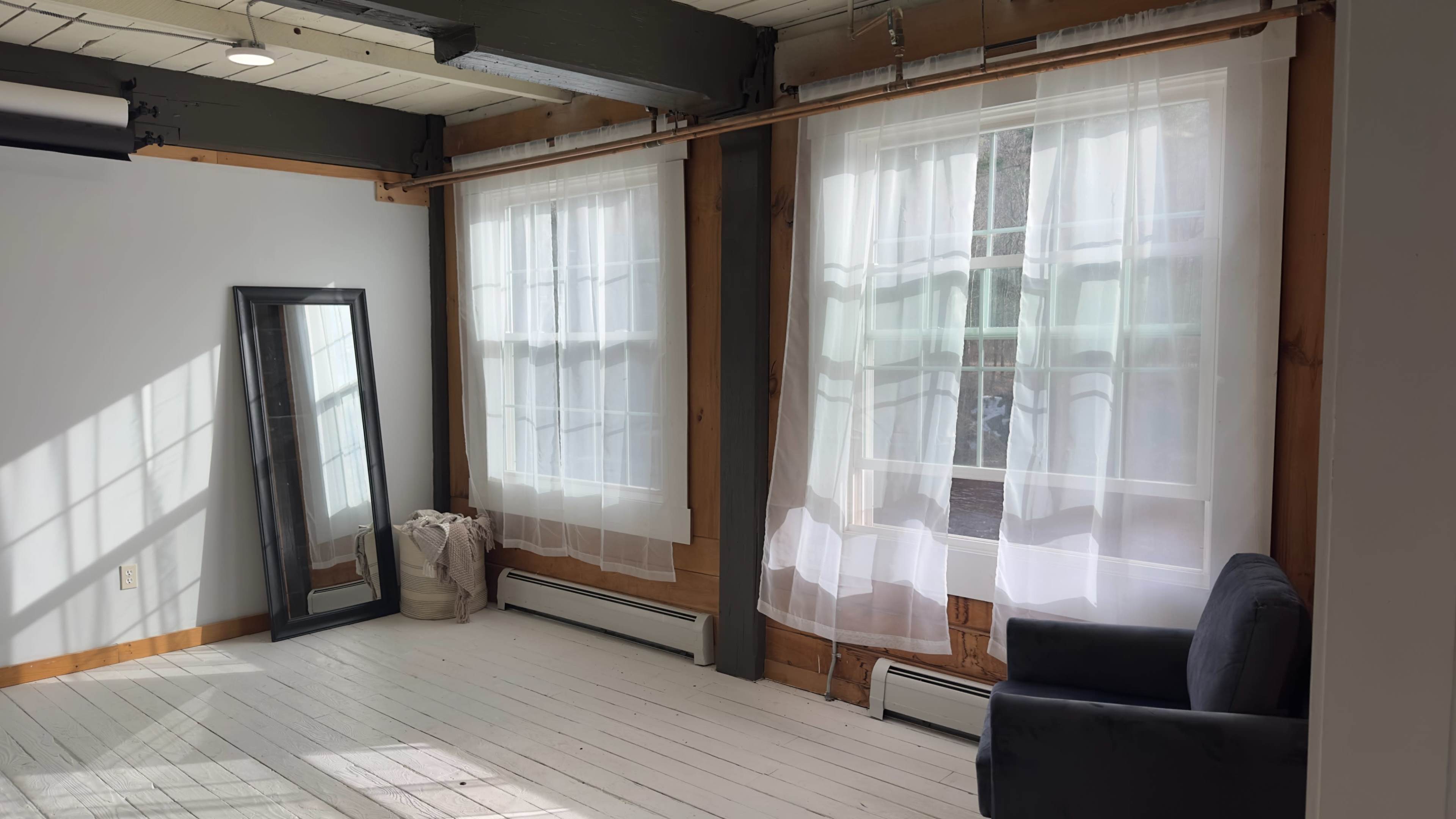 The image shows a well-lit room featuring two large windows with sheer curtains, a mirror against one wall, and a dark sofa.