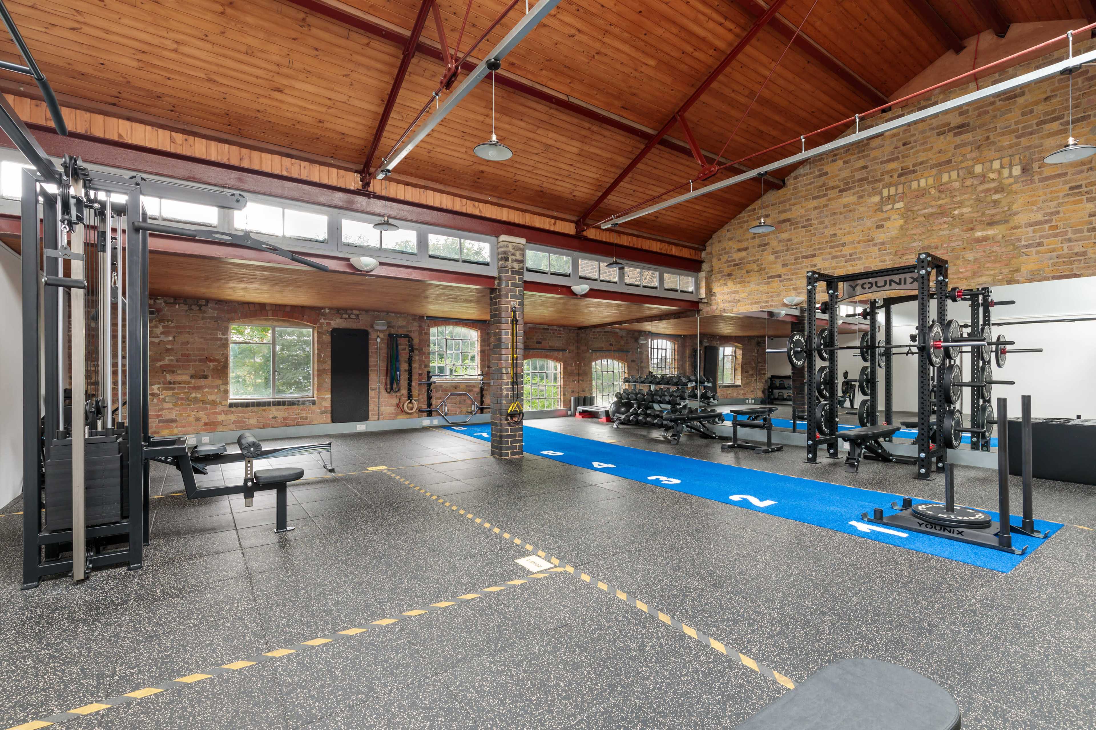 A spacious gym with various weightlifting equipment, rubber flooring, and large windows that allow natural light to enter.