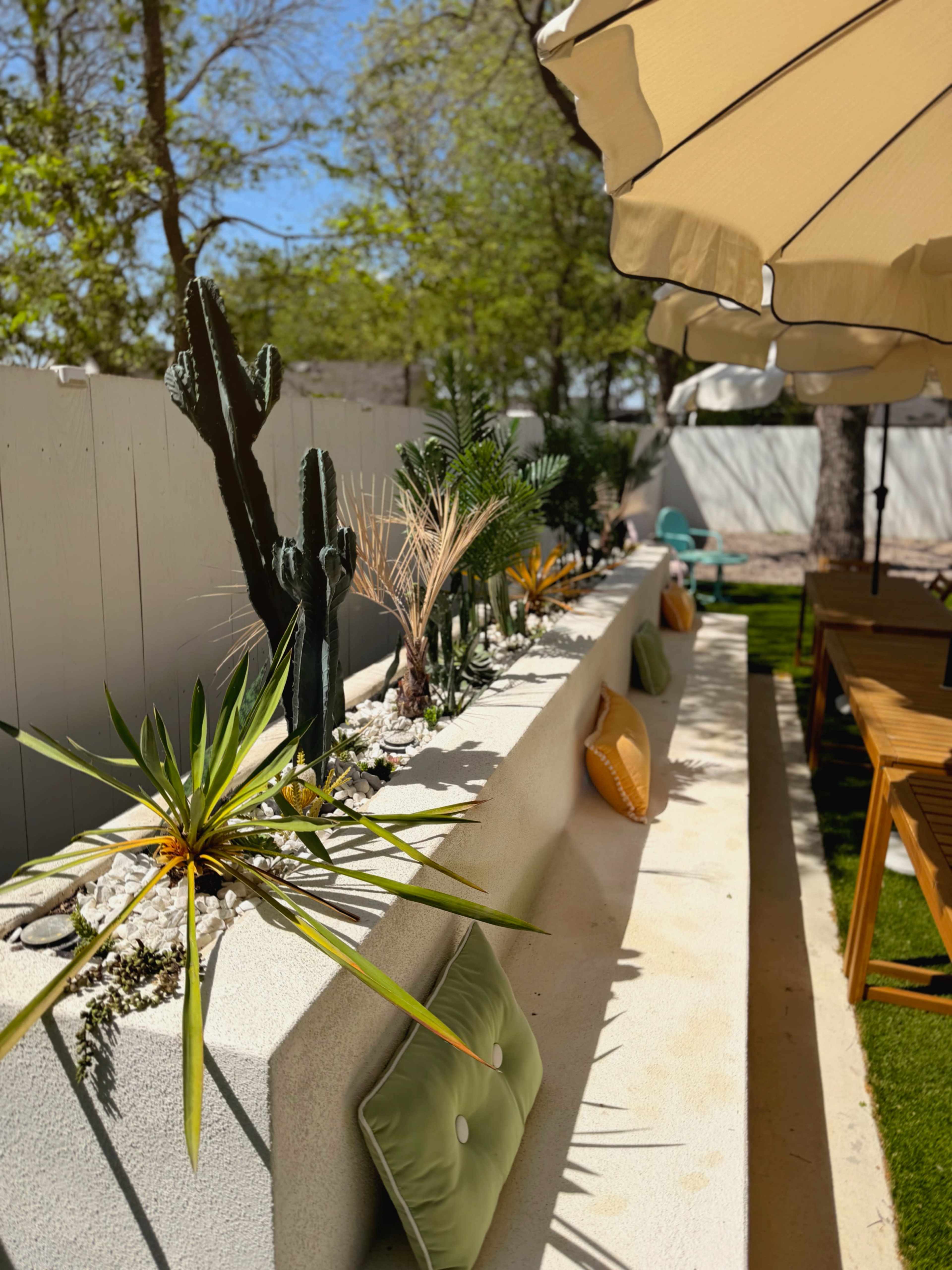 A concrete planter is arranged with various plants and decorative cushions, alongside a sun umbrella and outdoor seating in a landscaped area.