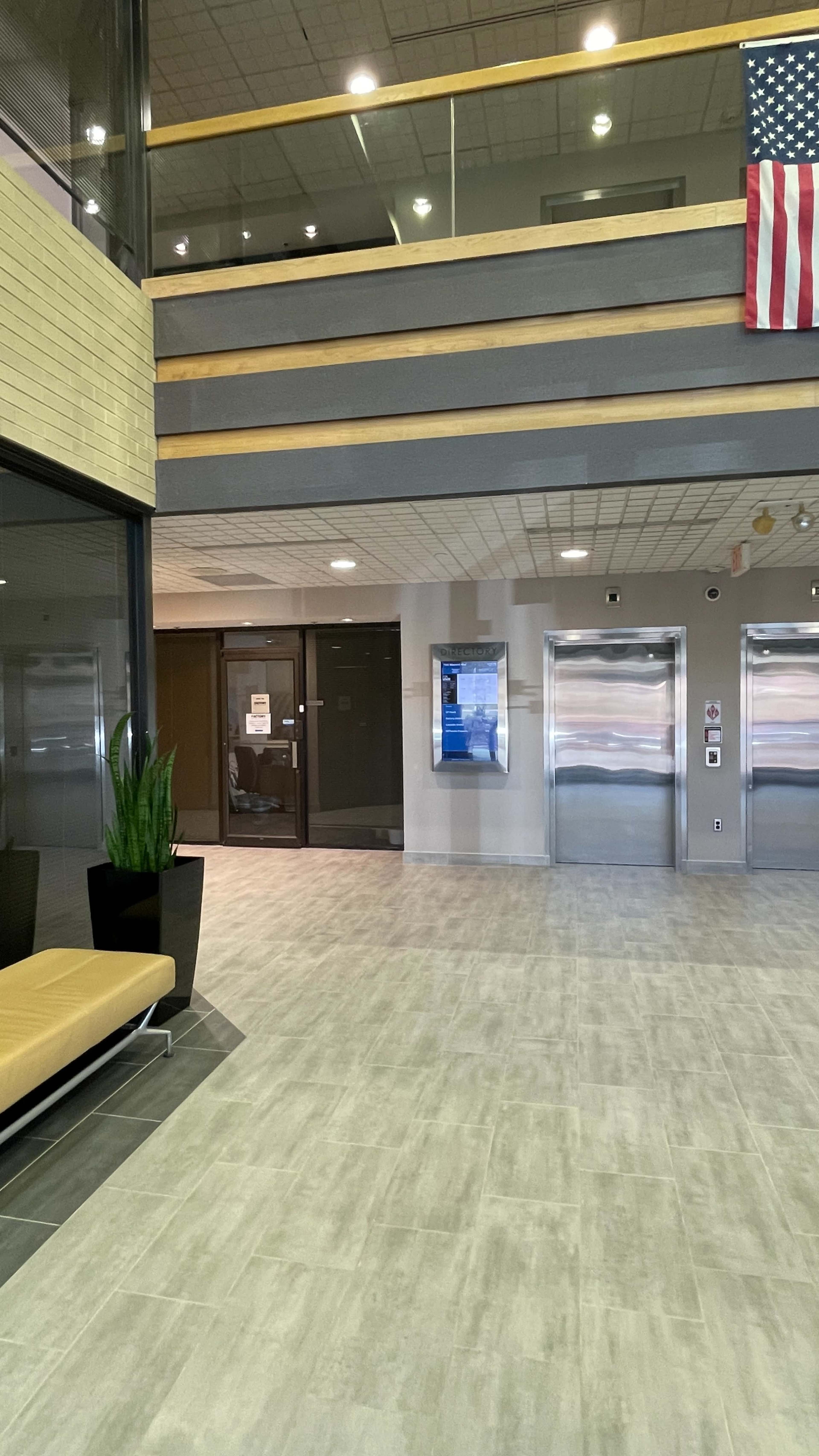 The image shows a modern lobby area with elevators, a seating bench, and an American flag displayed on the wall.