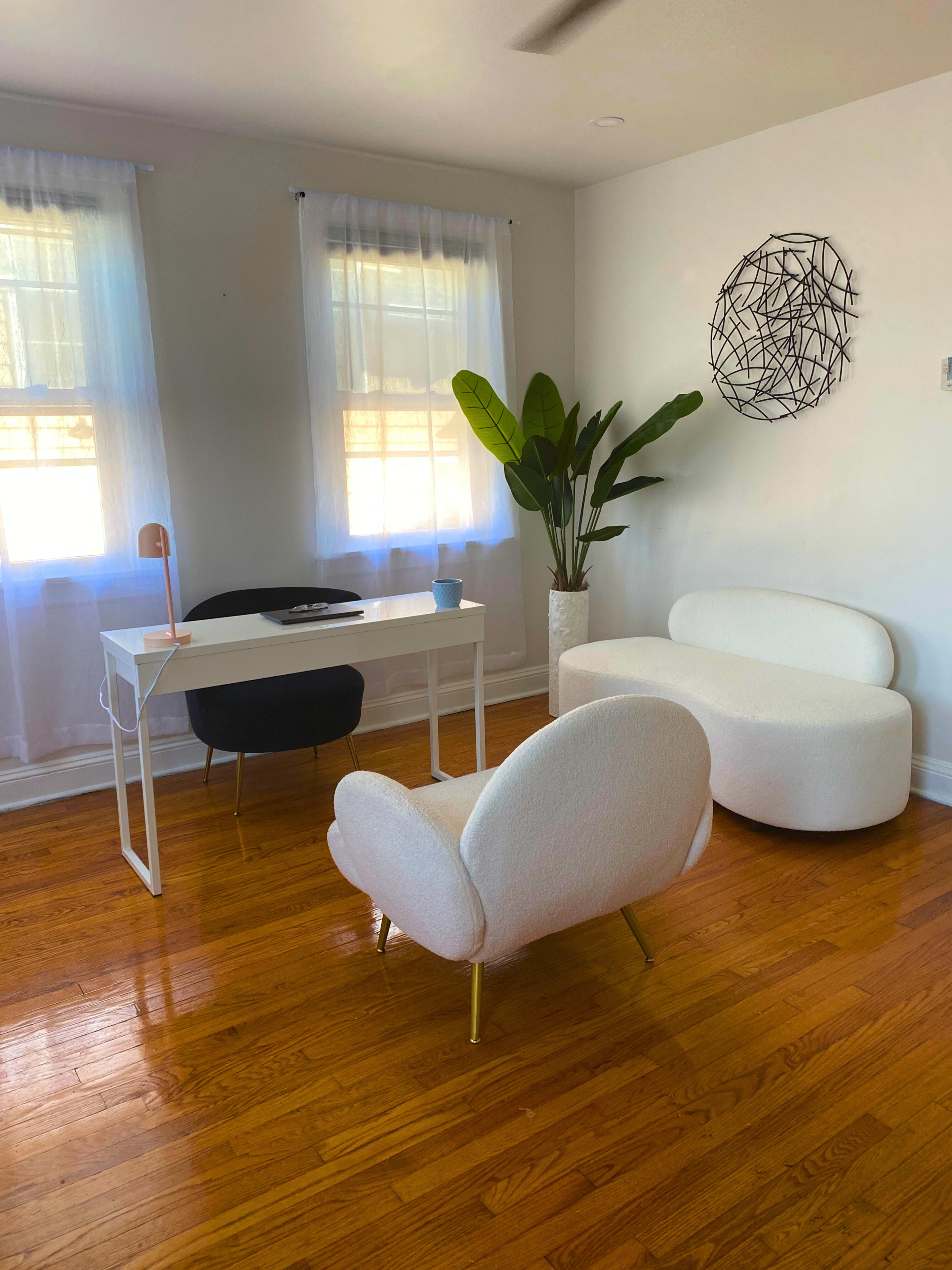 A bright, minimalistic office space with a white desk, a black chair, a white sculptural chair, a small table, and a large potted plant.