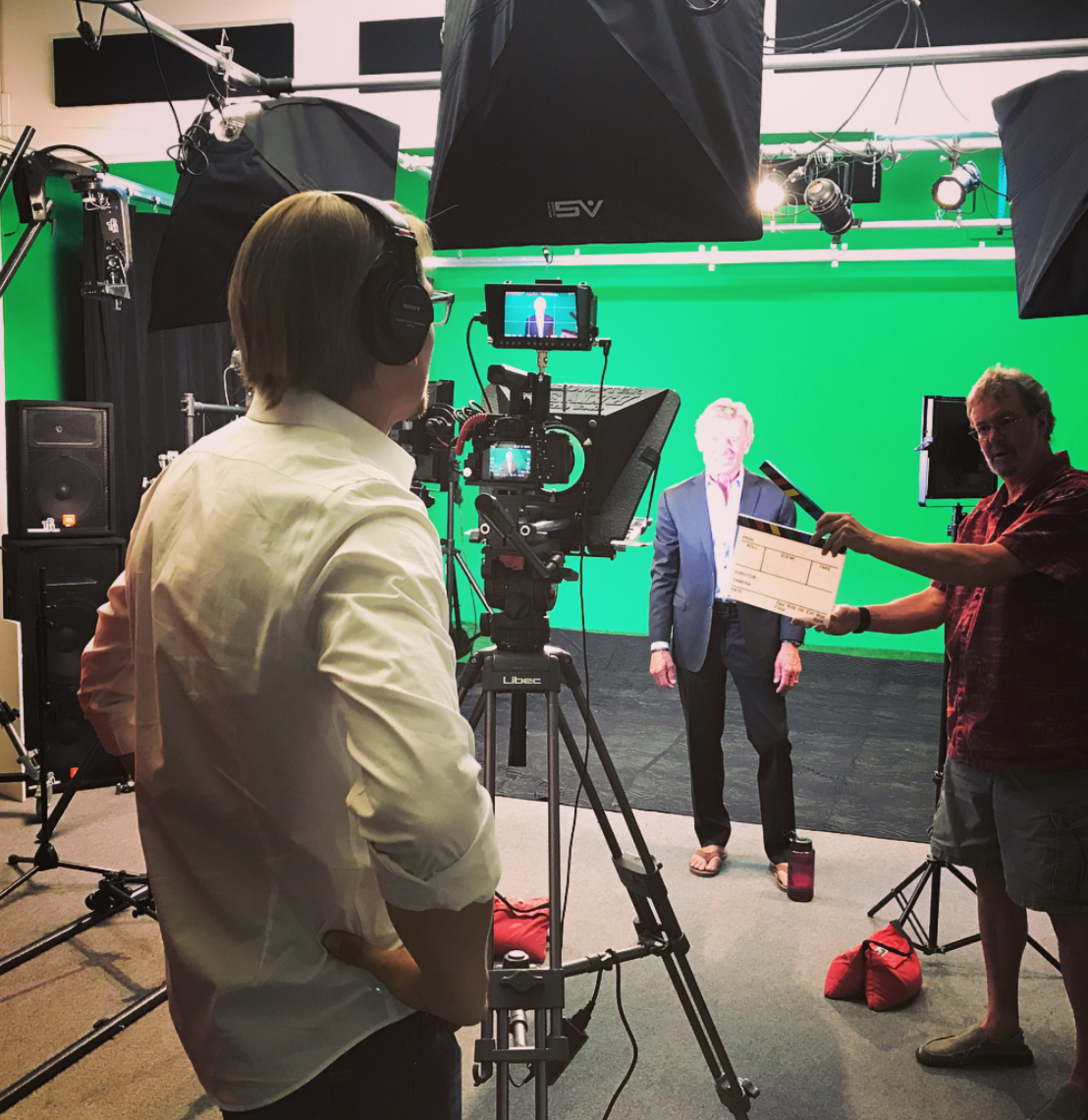 A man in a suit stands in front of a green screen while another man holds a clapperboard and operates a camera.