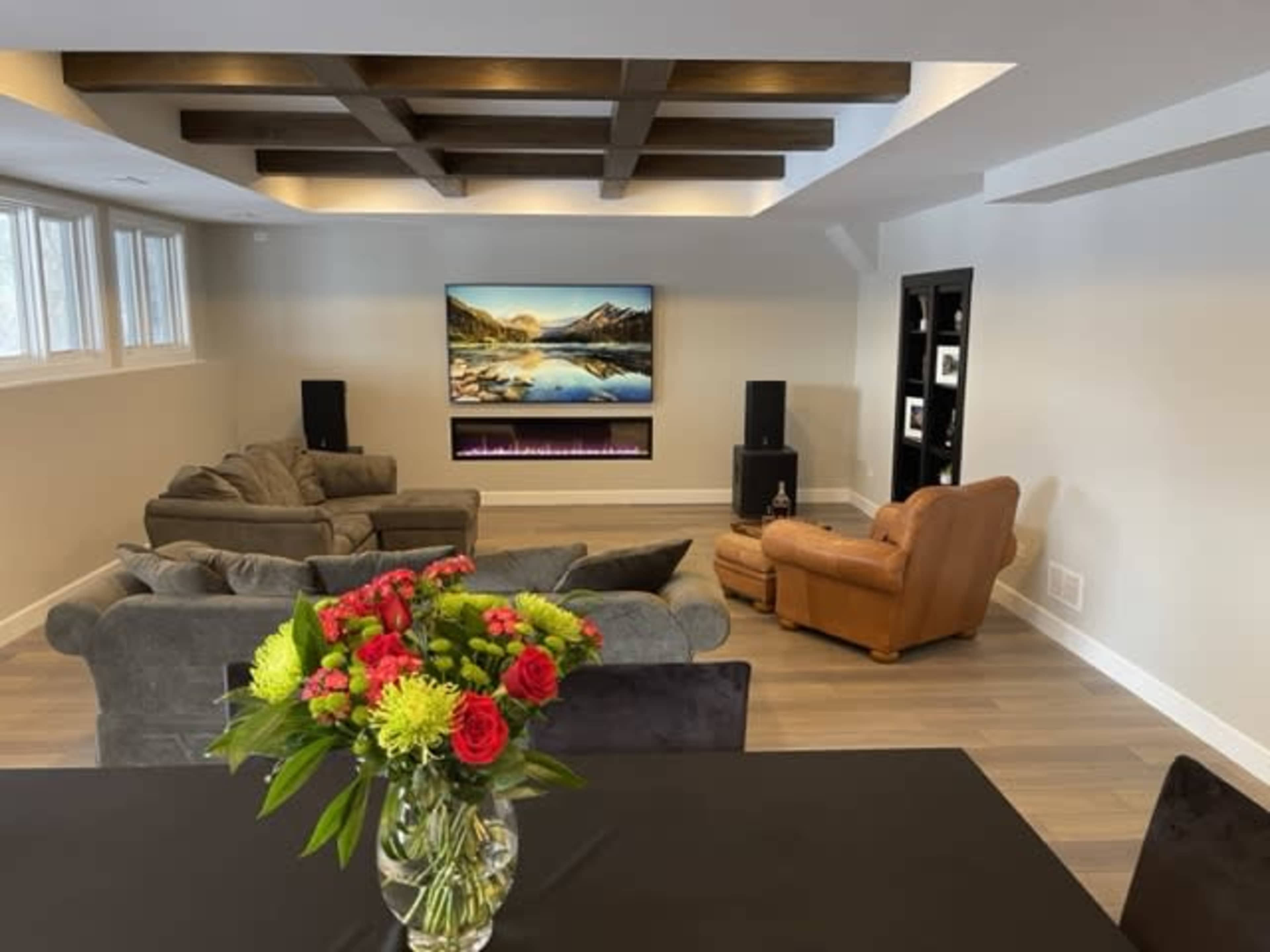 A modern living room features a large television mounted on the wall, a sofa set, a recliner, and a dining table with a floral centerpiece.