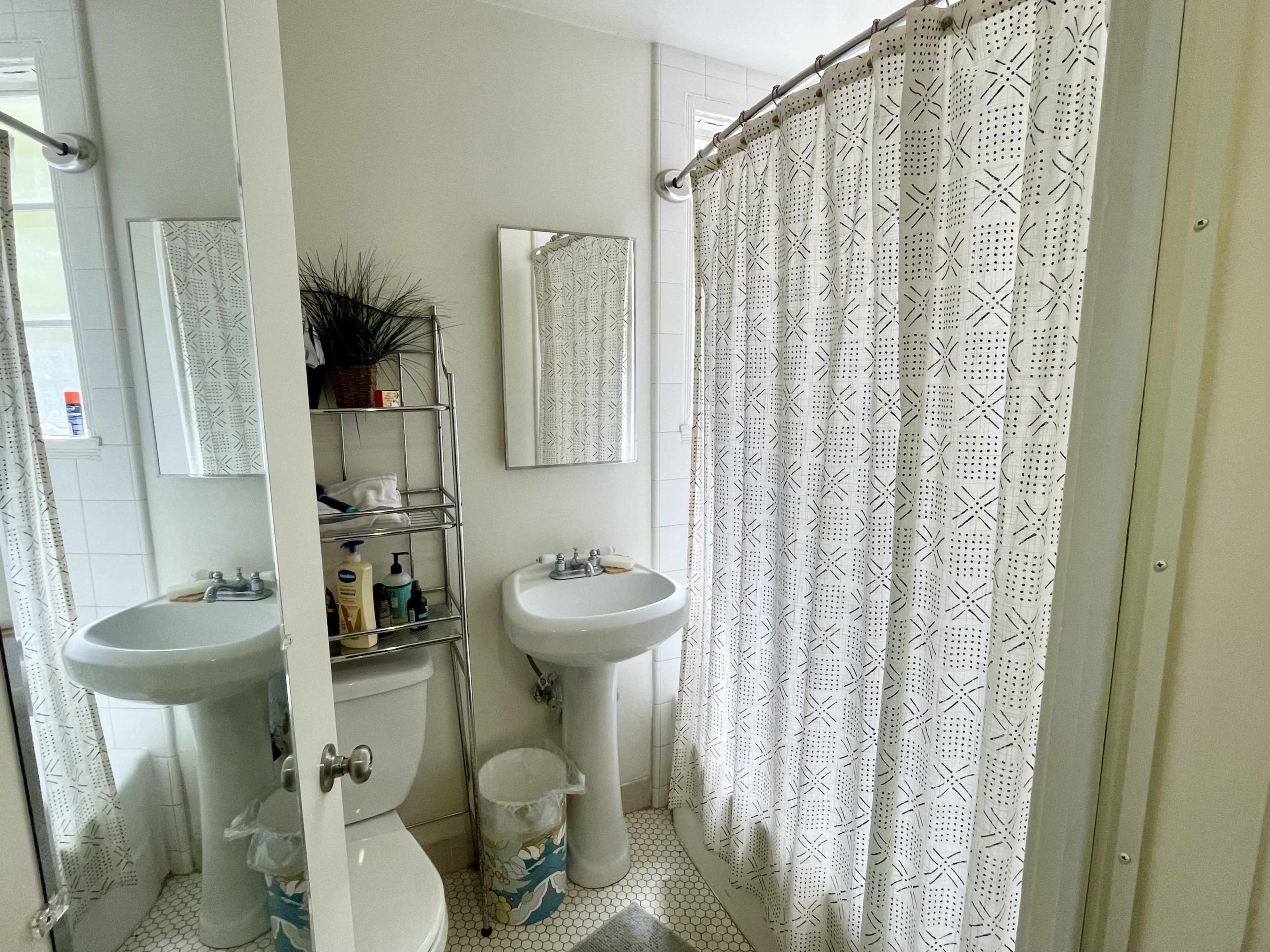 The image shows a small bathroom featuring two pedestal sinks, a shower with a patterned curtain, a metal shelving unit with toiletries, and a toilet.