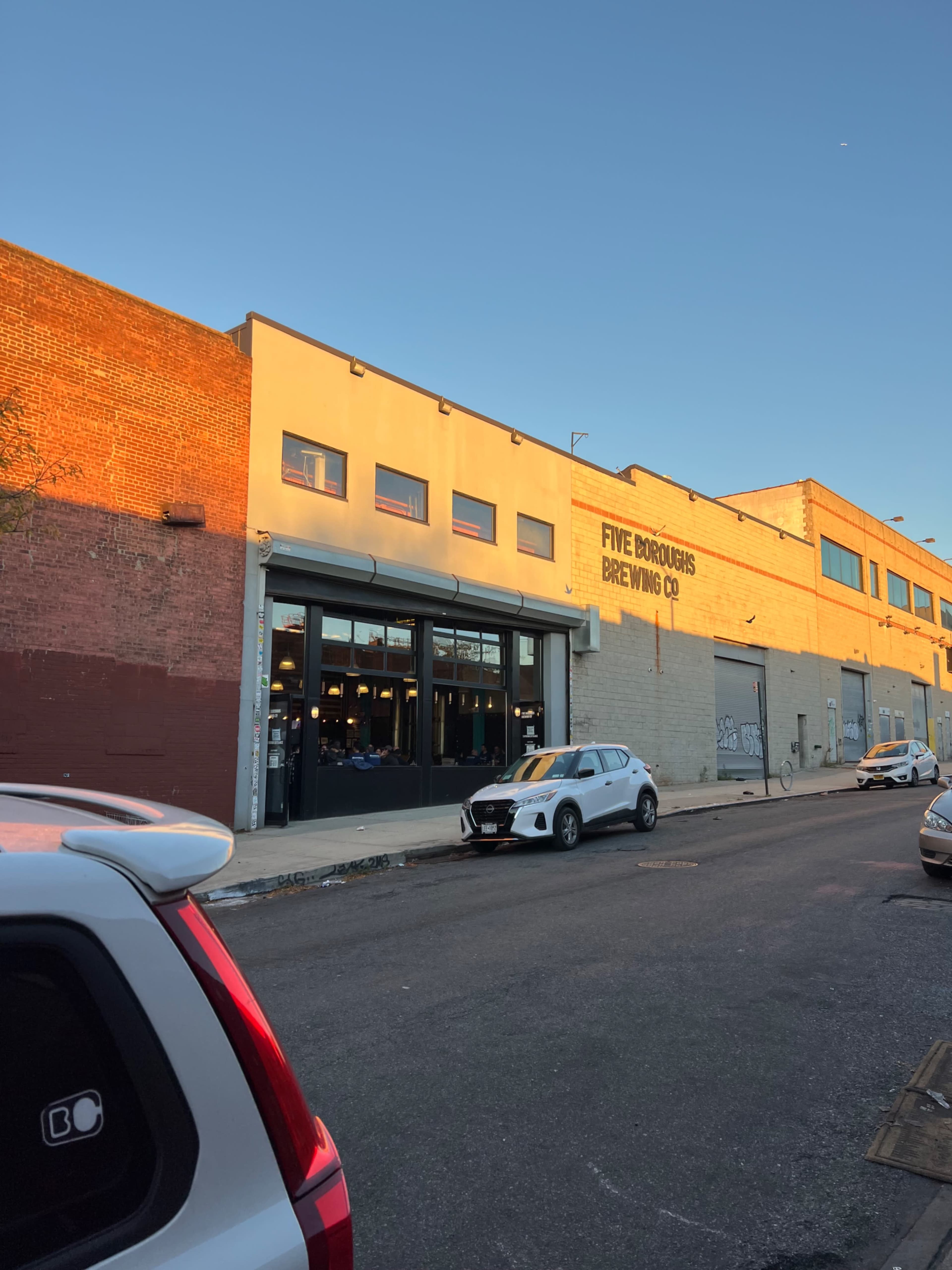 The image shows a brewing facility named "Five Boroughs Brewing Co." with a clear blue sky overhead and parked cars in front.