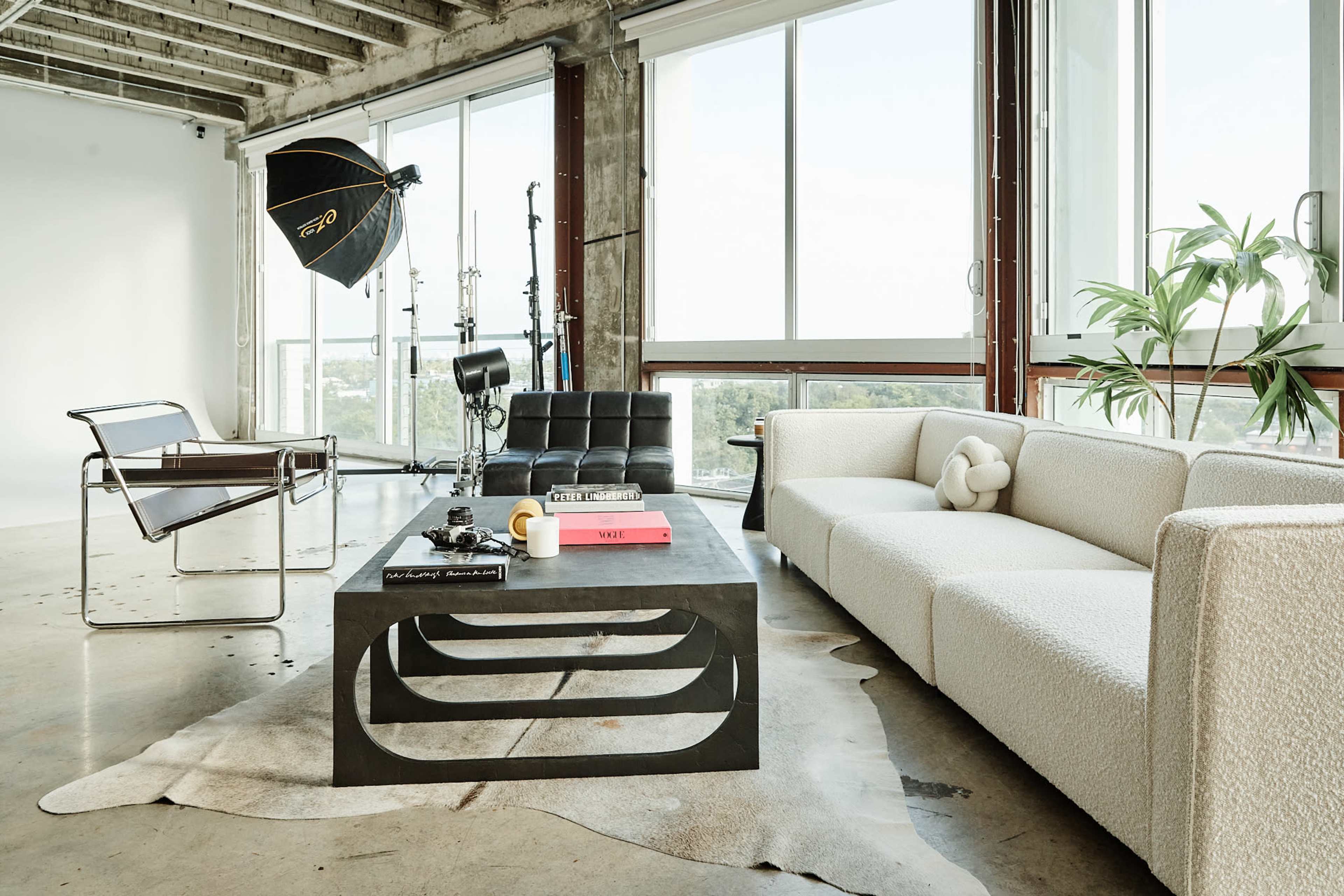 A modern studio space with a large window, showcasing a minimalist seating area that includes a white sofa, a black chair, and a coffee table with various items on it.