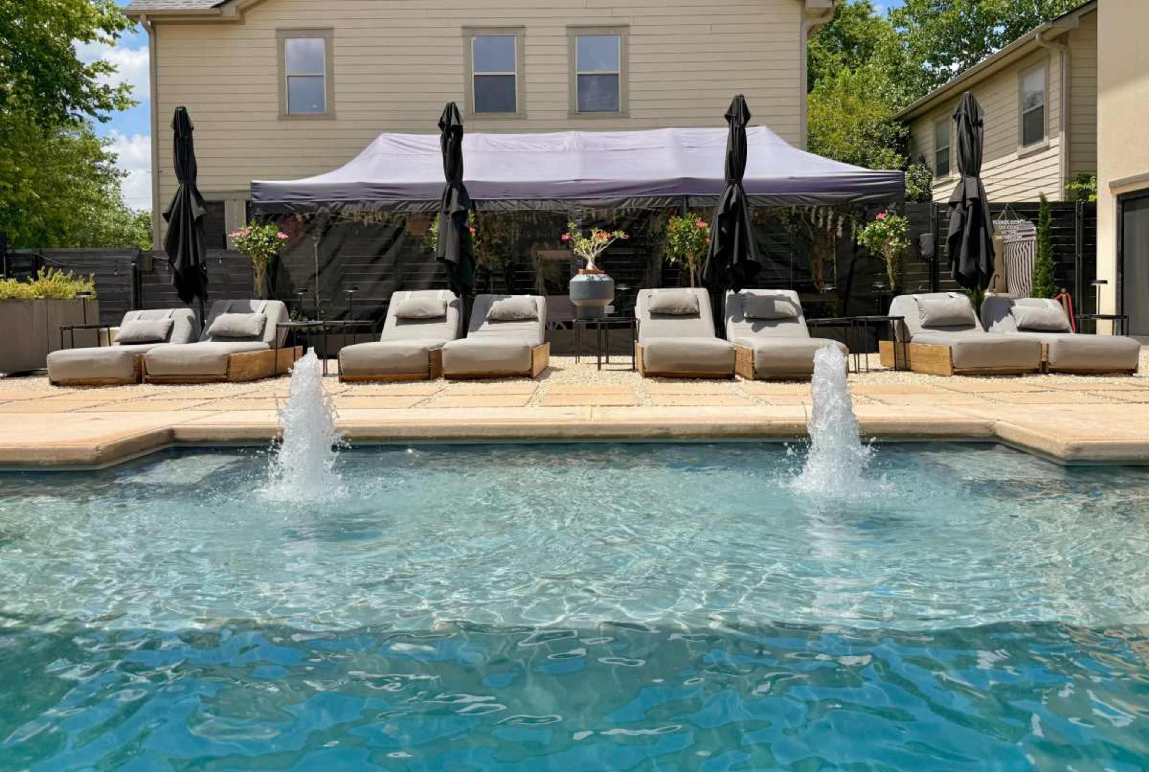 A swimming pool with two fountains in the foreground is bordered by lounge chairs and a shaded seating area with black umbrellas in the background.