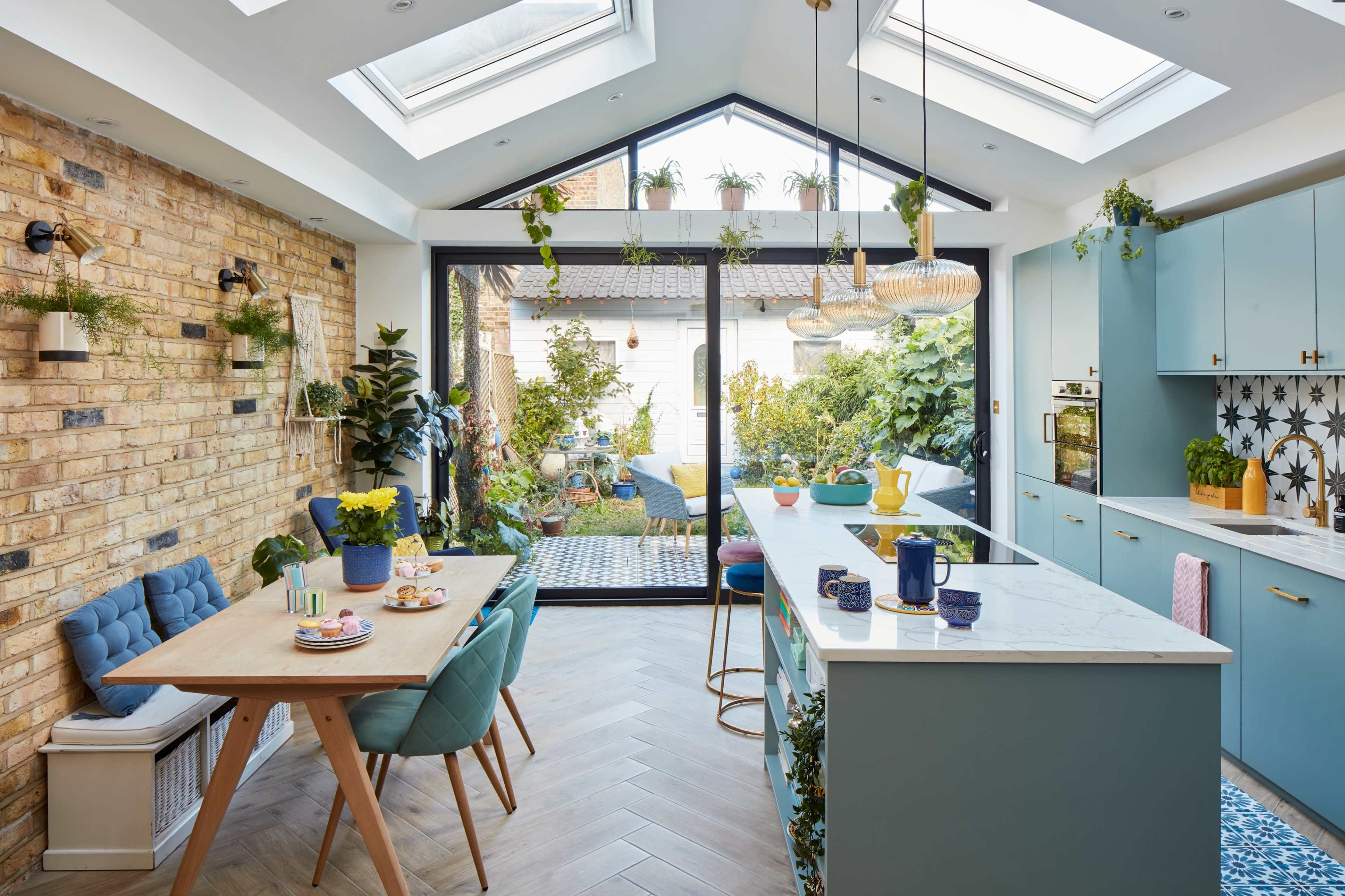 A modern kitchen with a large island, blue cabinetry, a dining table set for a meal, and abundant greenery seen through large glass doors.
