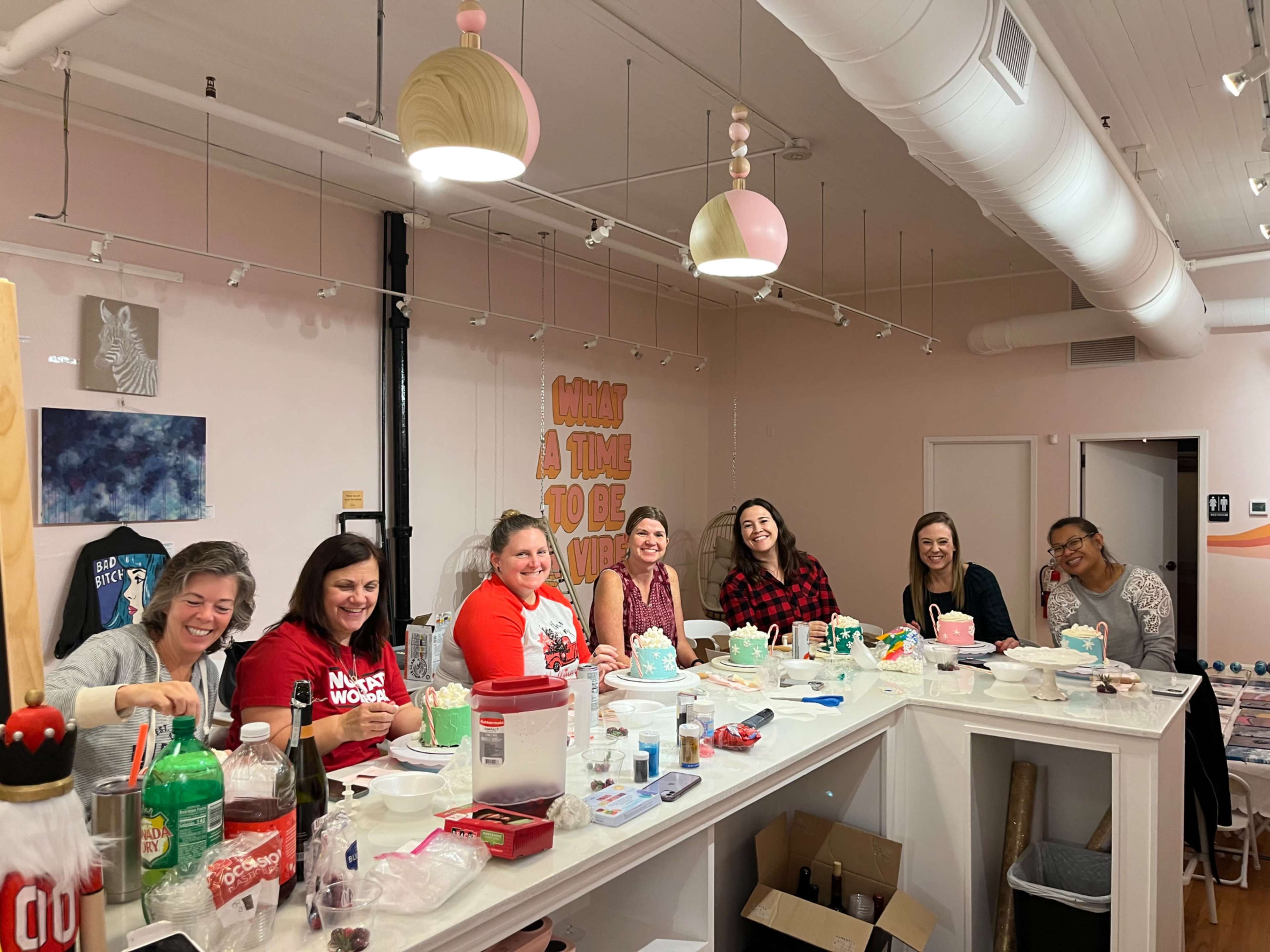 A group of eight women are engaged in a crafting activity at a table, surrounded by supplies and decorated with pastel-colored hanging lamps.