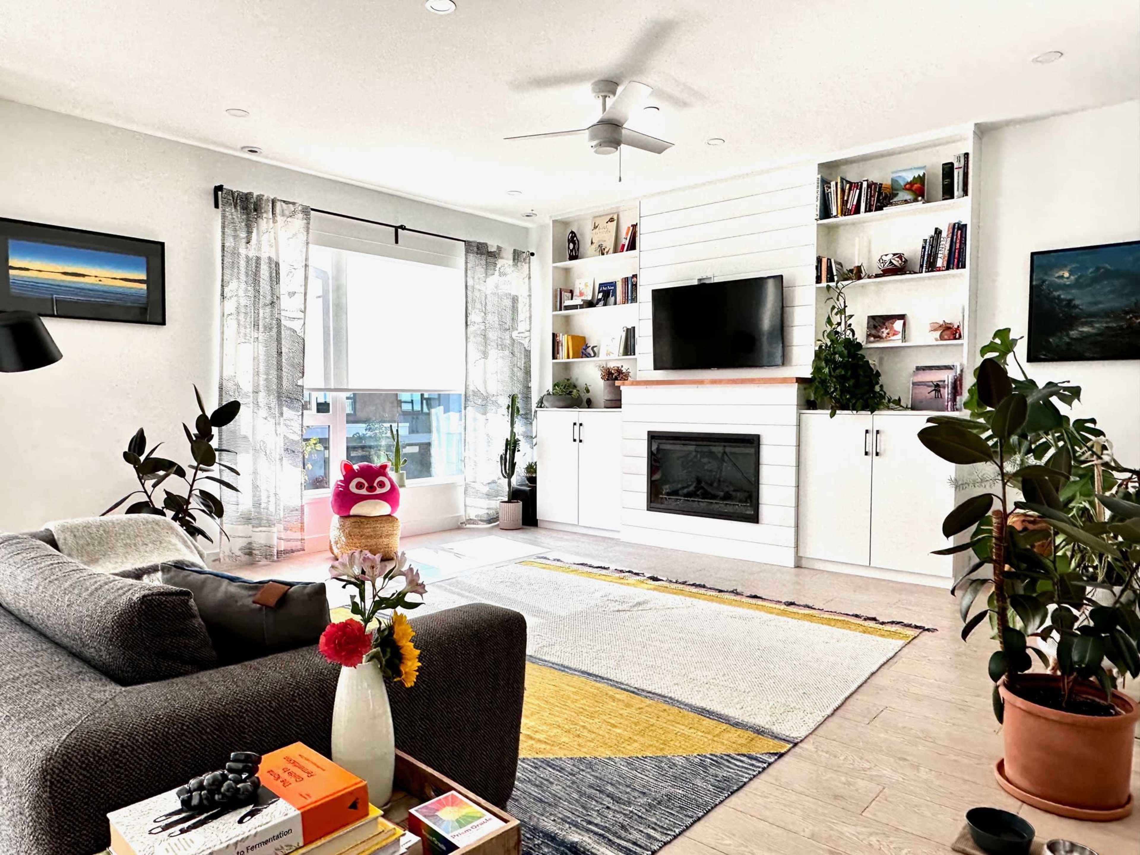 A modern living room features a gray sofa, a television mounted above a fireplace, shelves filled with books and decor, and large windows with natural light.