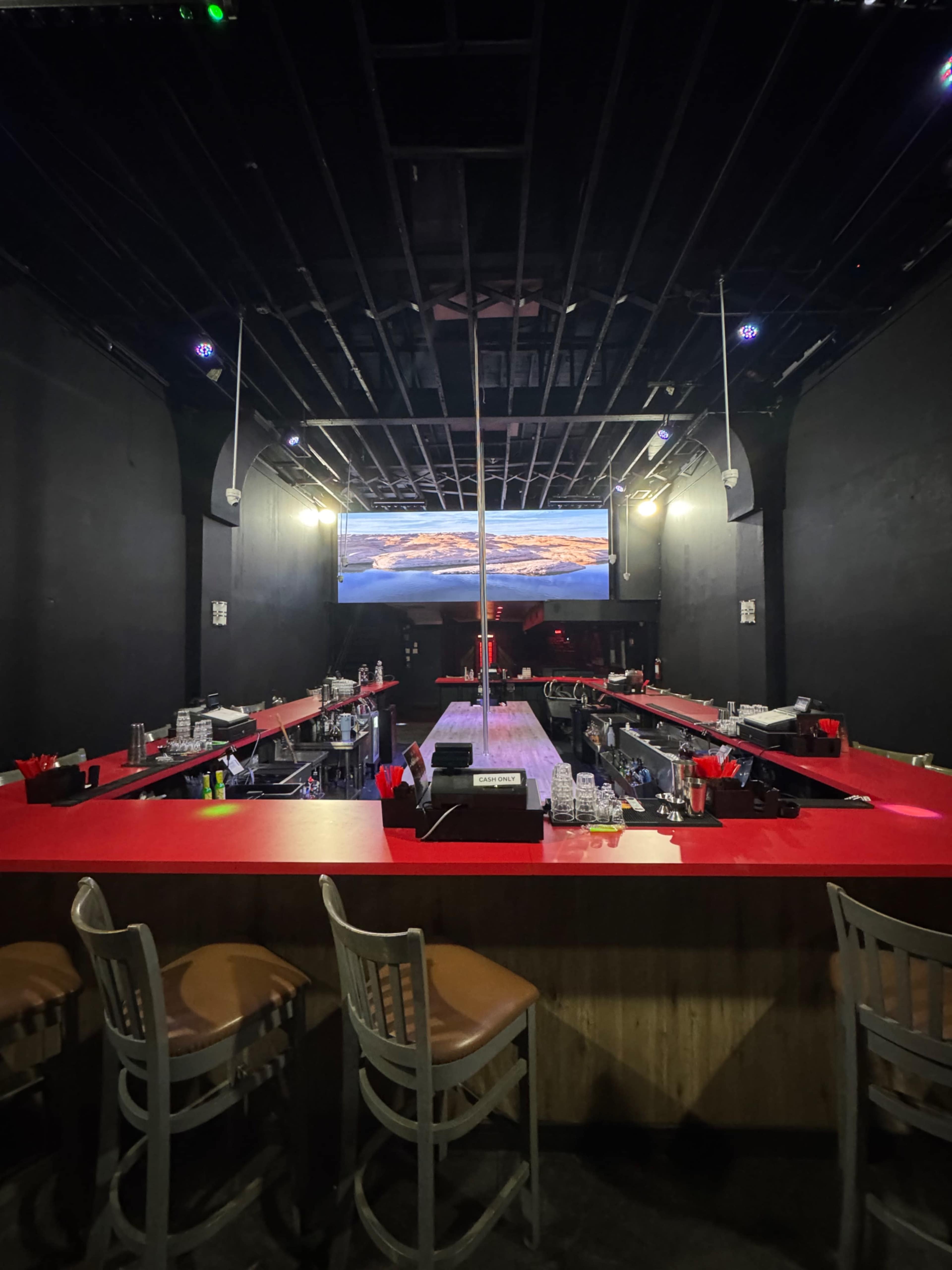 The image shows a dimly lit bar with red countertops and wooden stools, featuring a long central area and a large screen at the back displaying a landscape.