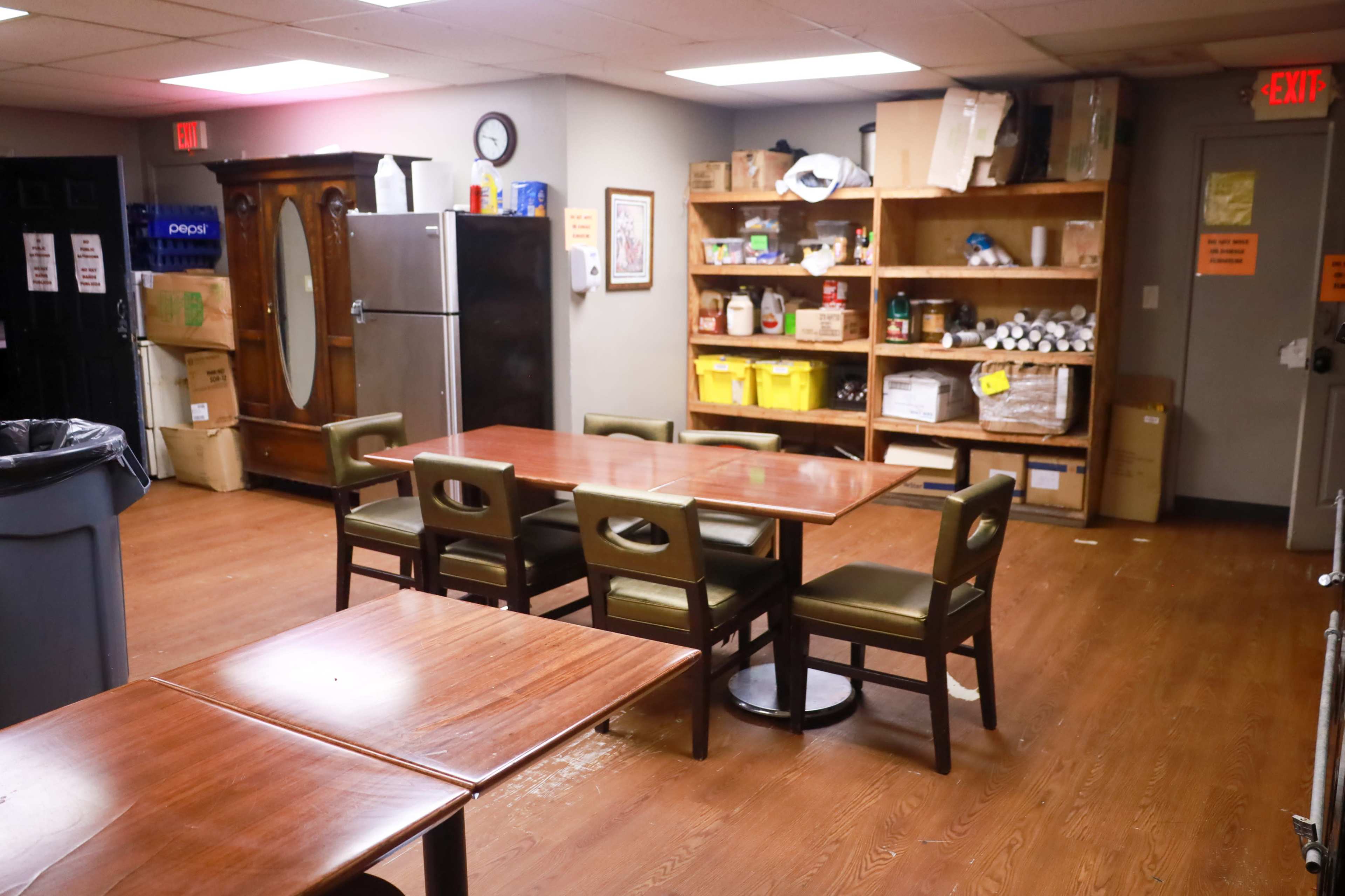 The image depicts a sparsely furnished break room with a table and chairs, a refrigerator, and shelves filled with assorted food items and supplies.