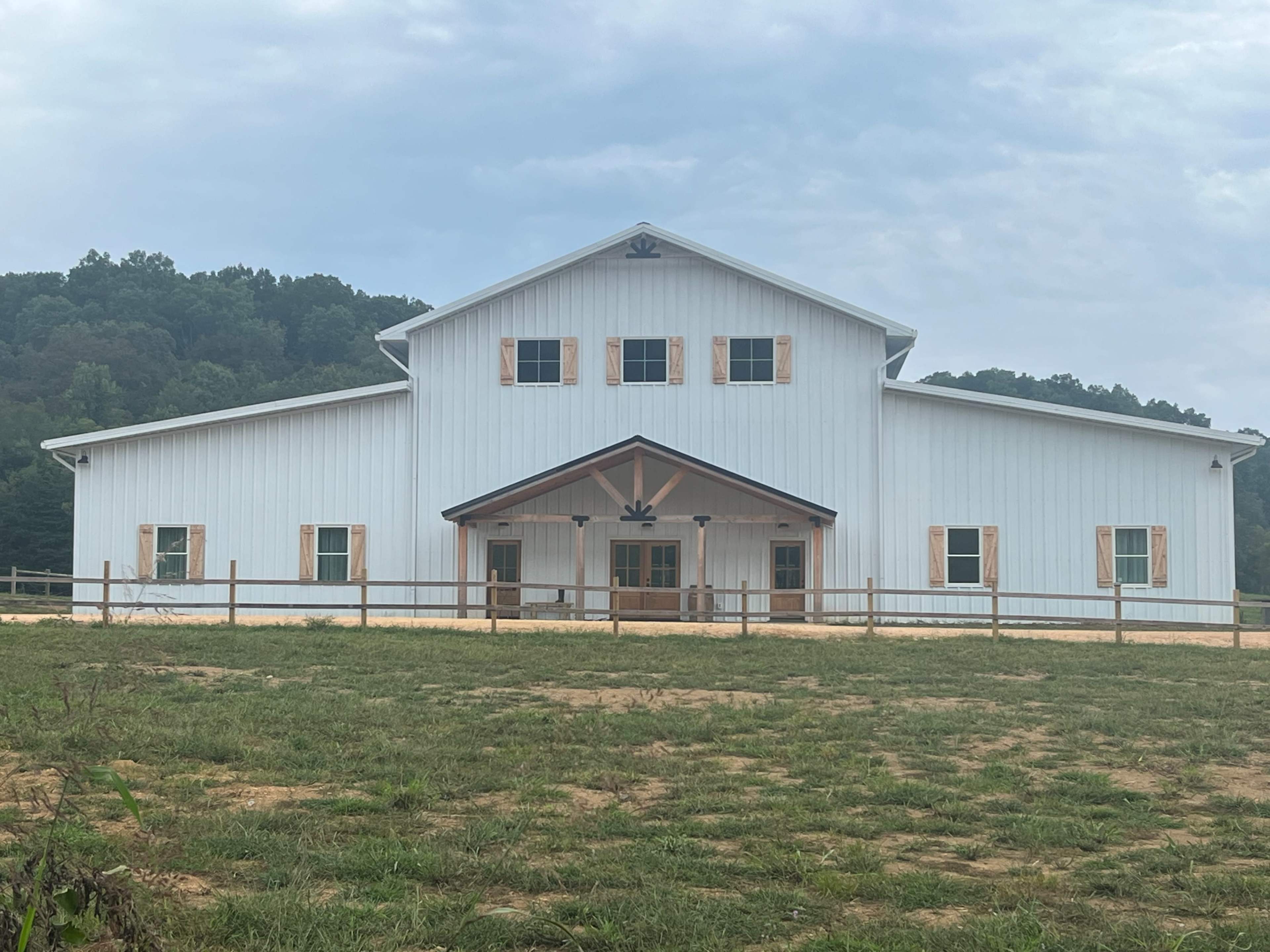 A large, white agricultural building with a steep roof and wooden shutters, surrounded by an open grassy area and trees in the background.