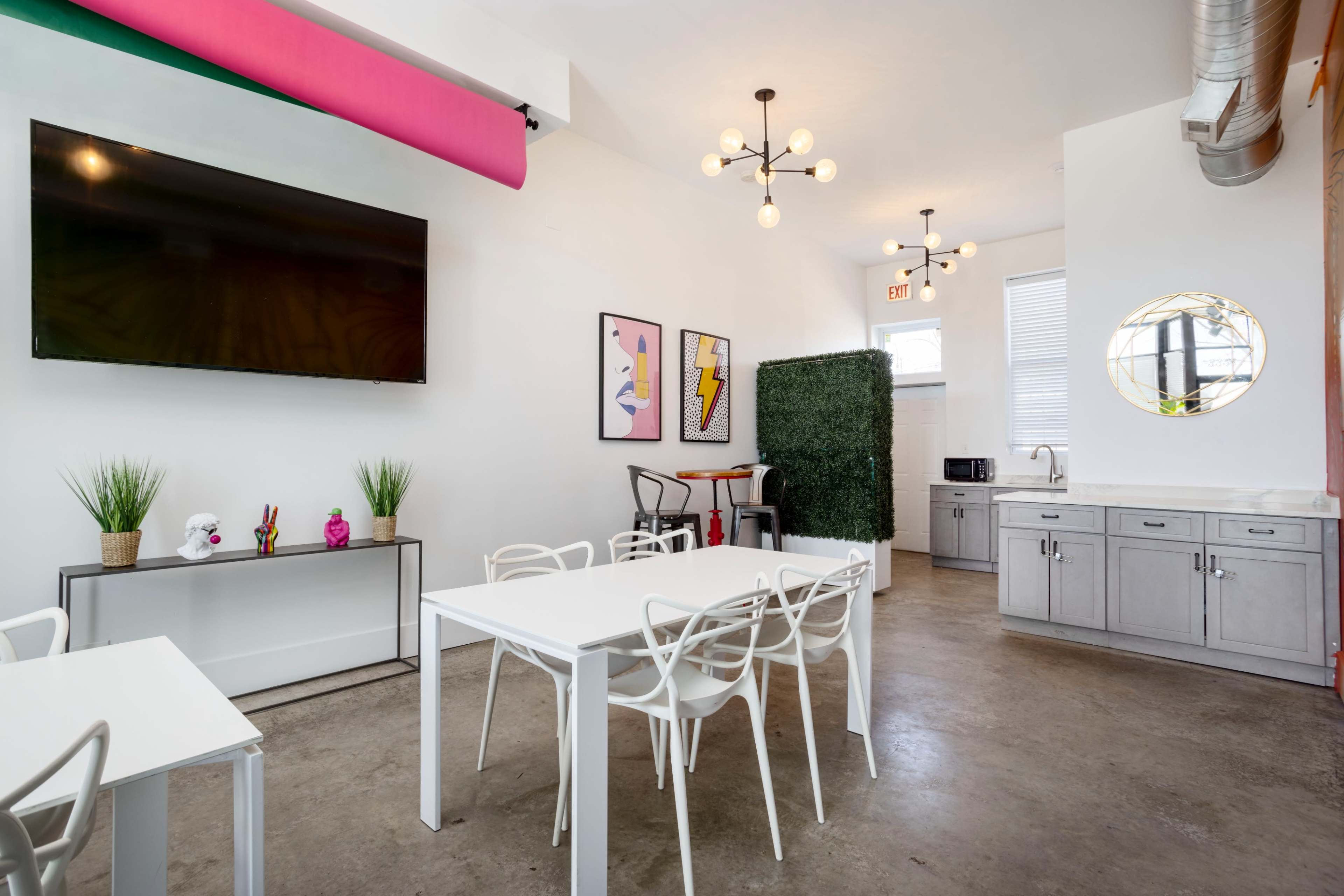 A modern, brightly lit room featuring white furniture, a large television on the wall, and a kitchenette area in the background.