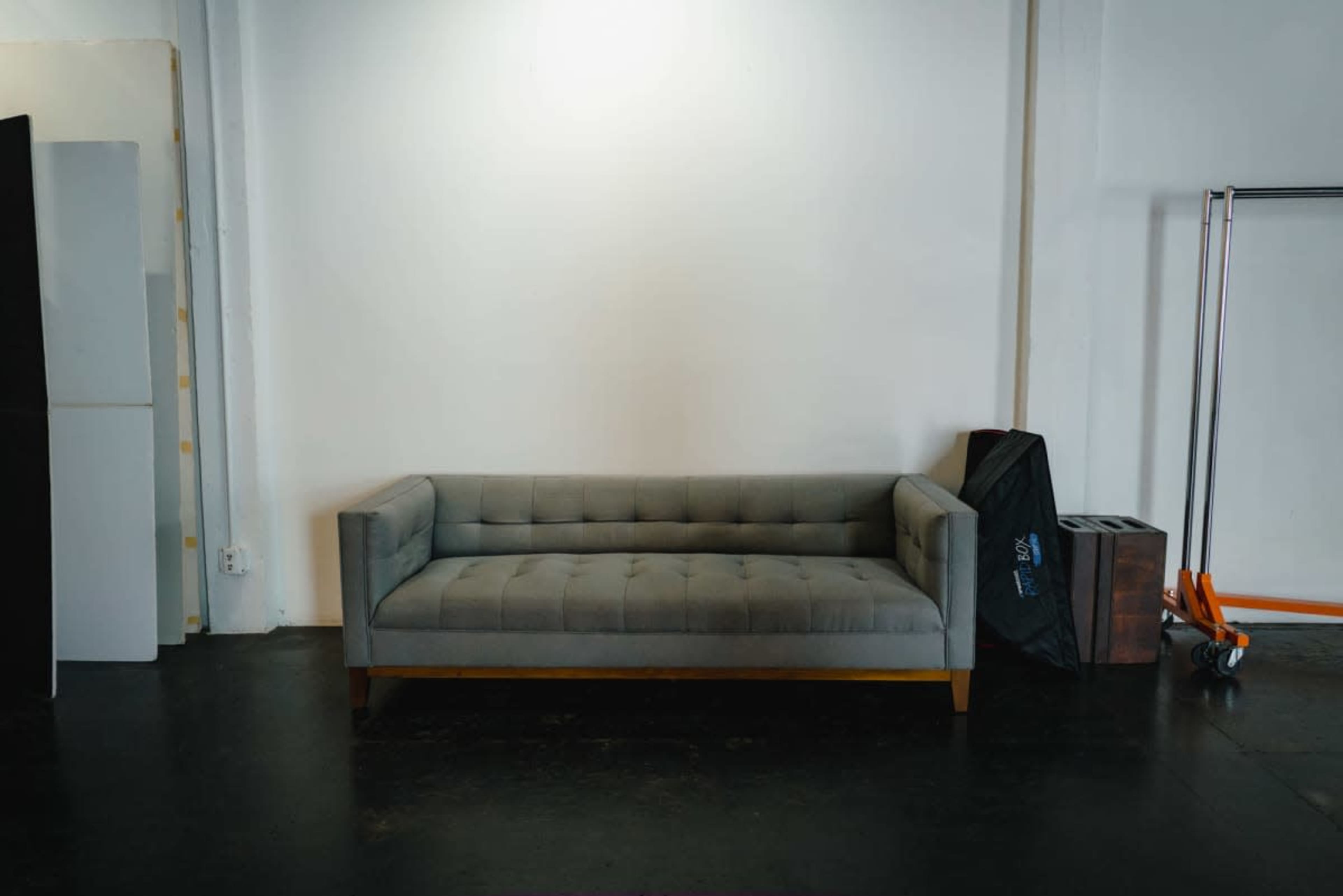 A gray tufted sofa is positioned against a white wall in a sparse, well-lit room with minimal furnishings.