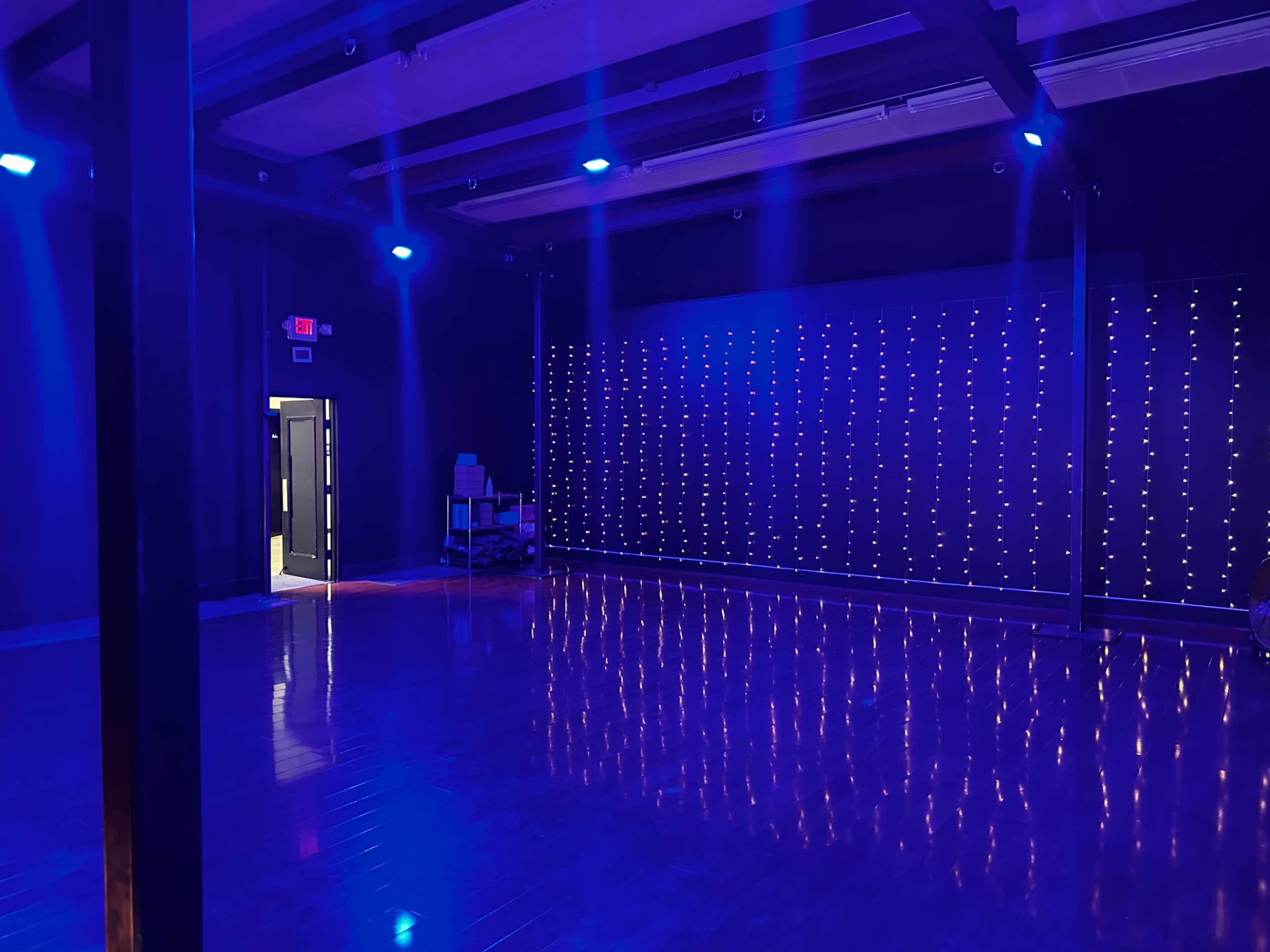 A dance studio with a polished wooden floor, a wall of blue lights, and an open doorway leading to another room.