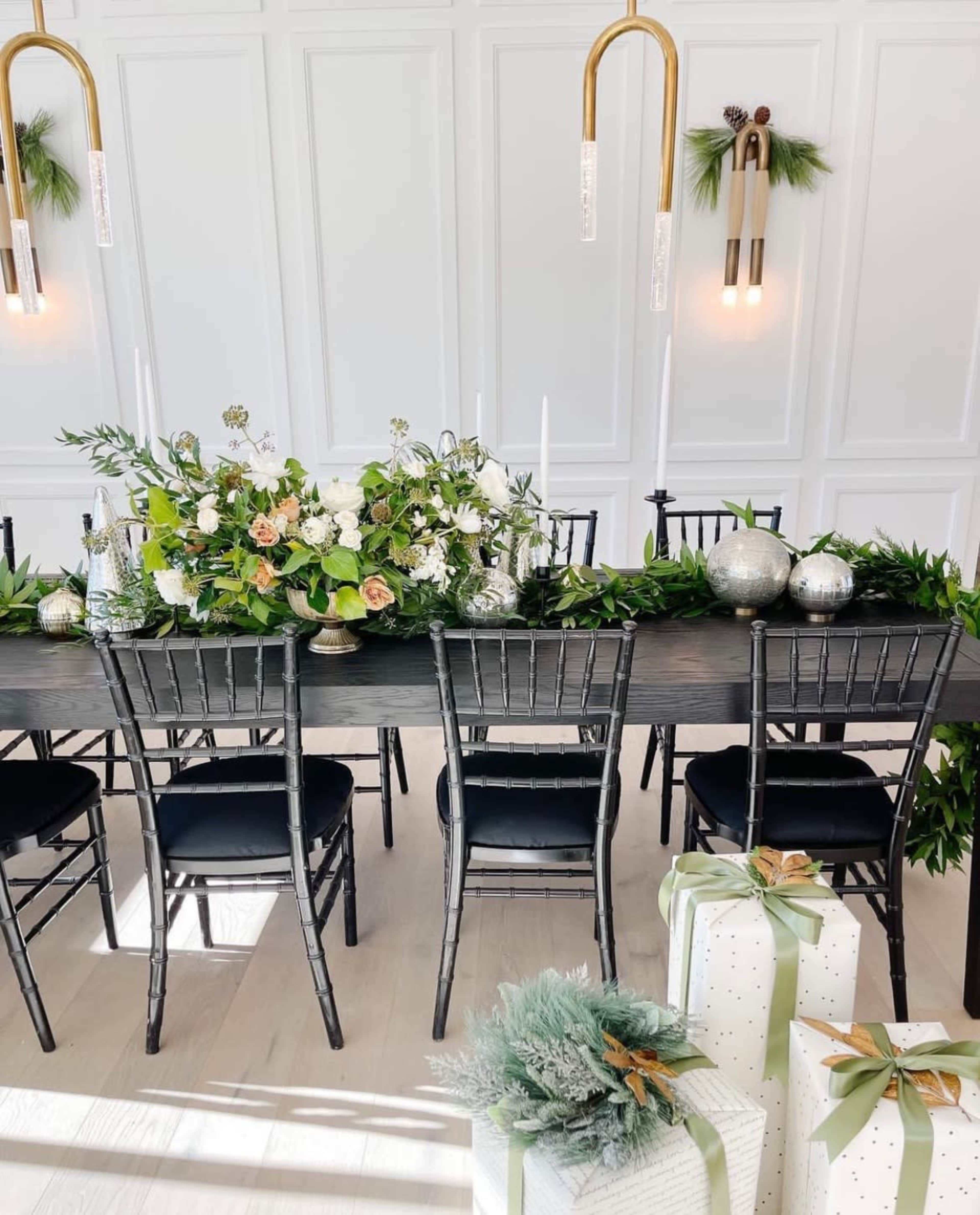 The image shows a long dining table decorated with floral arrangements, candles, and greenery, flanked by black chairs and adorned with wrapped gifts at the ends.