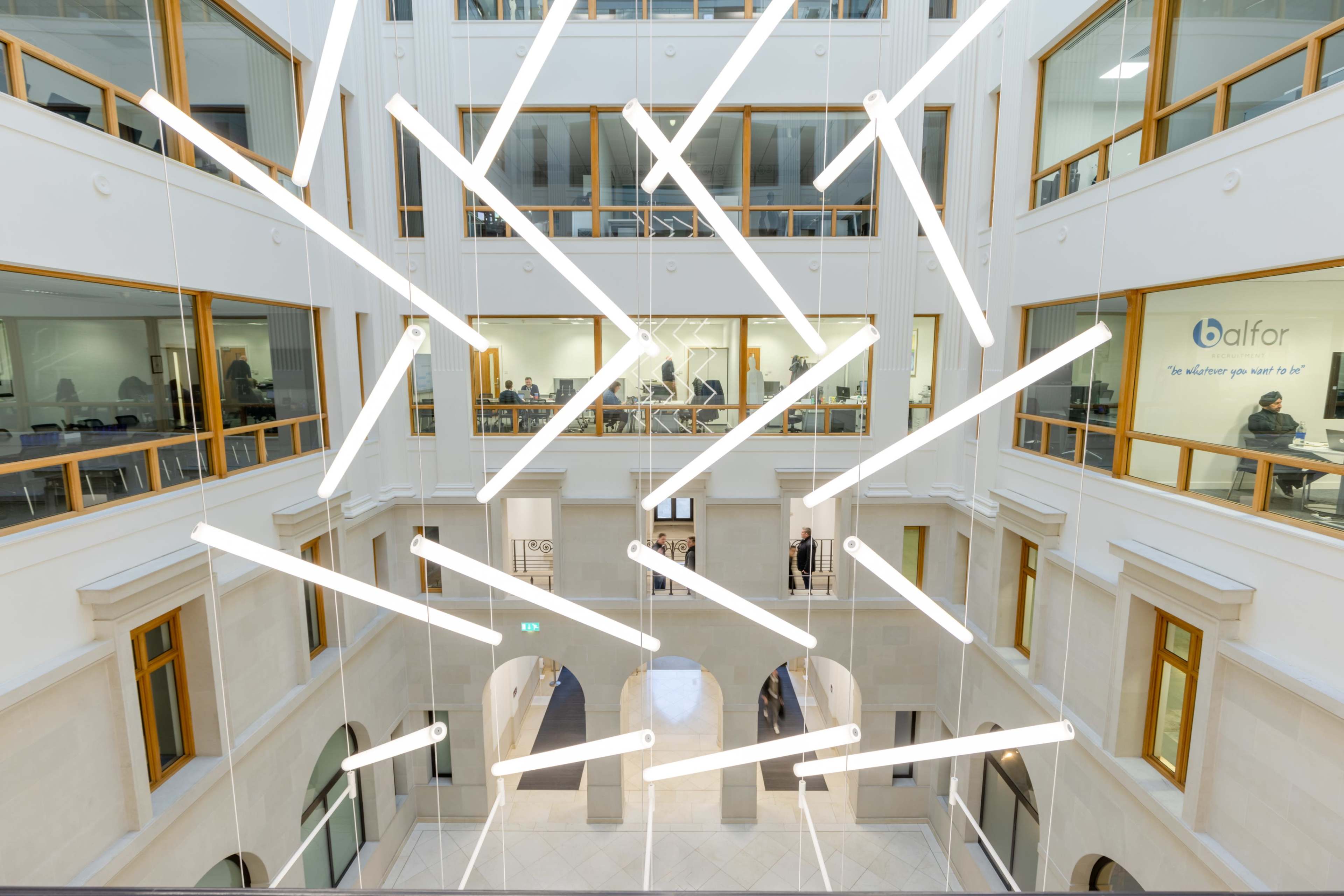 An interior view of a modern multi-level building with angular light fixtures suspended from the ceiling and several people visible in the background.