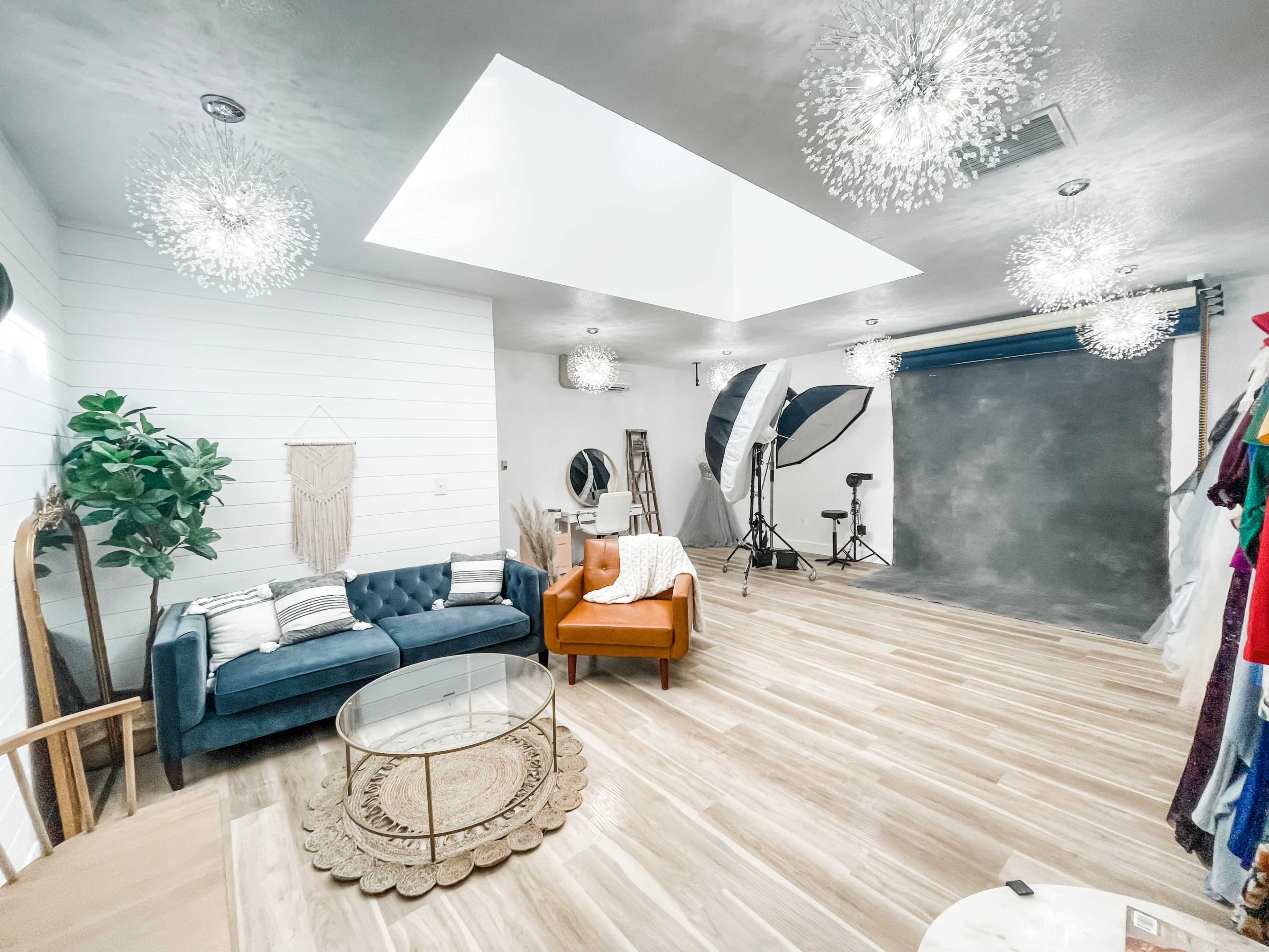 A spacious photography studio featuring a blue sofa, an orange armchair, a circular glass coffee table, and various lighting setups against a gray backdrop.
