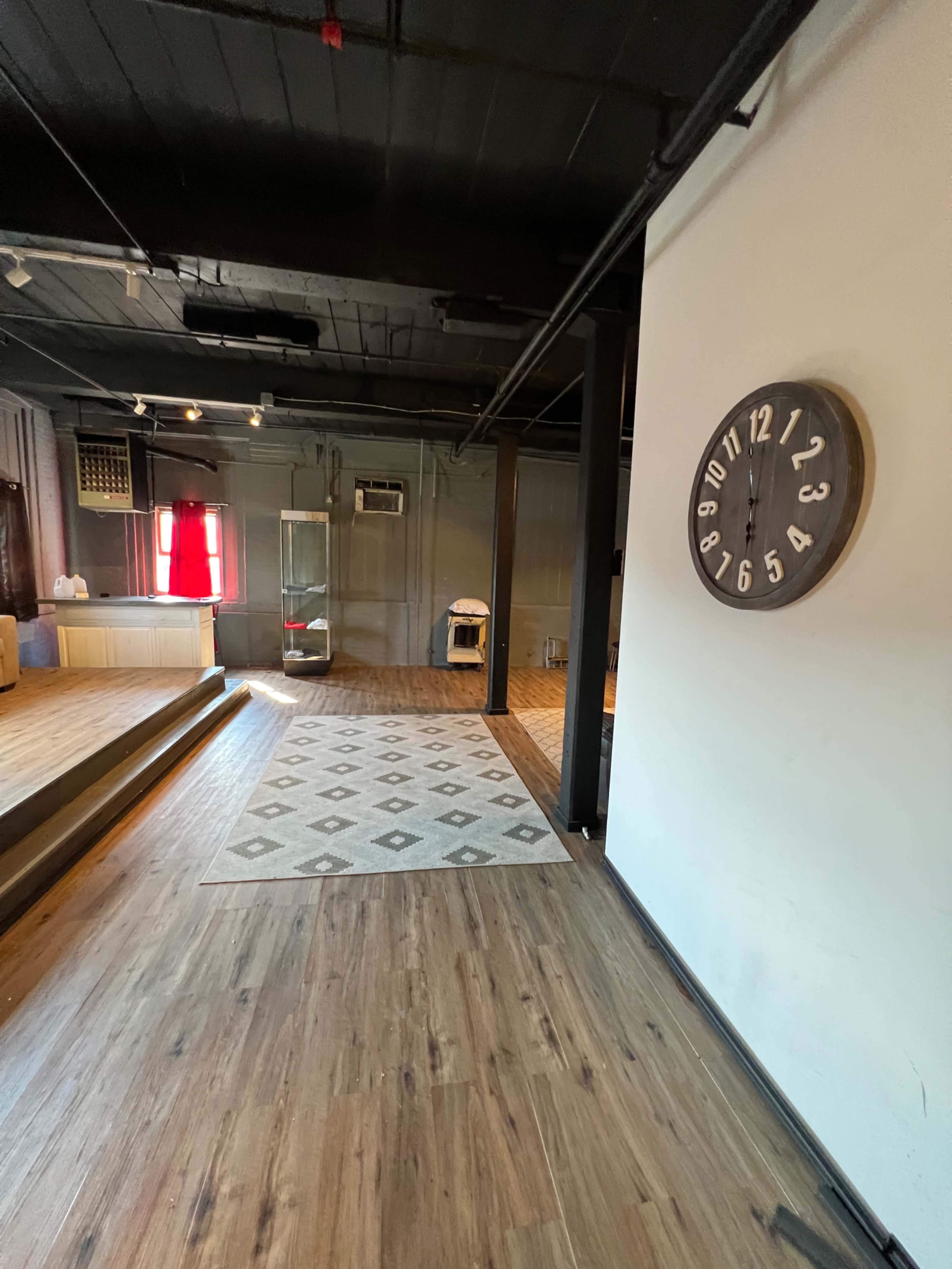 An interior space with wooden flooring, a wall clock, a patterned rug, and a small glass display case in the background.