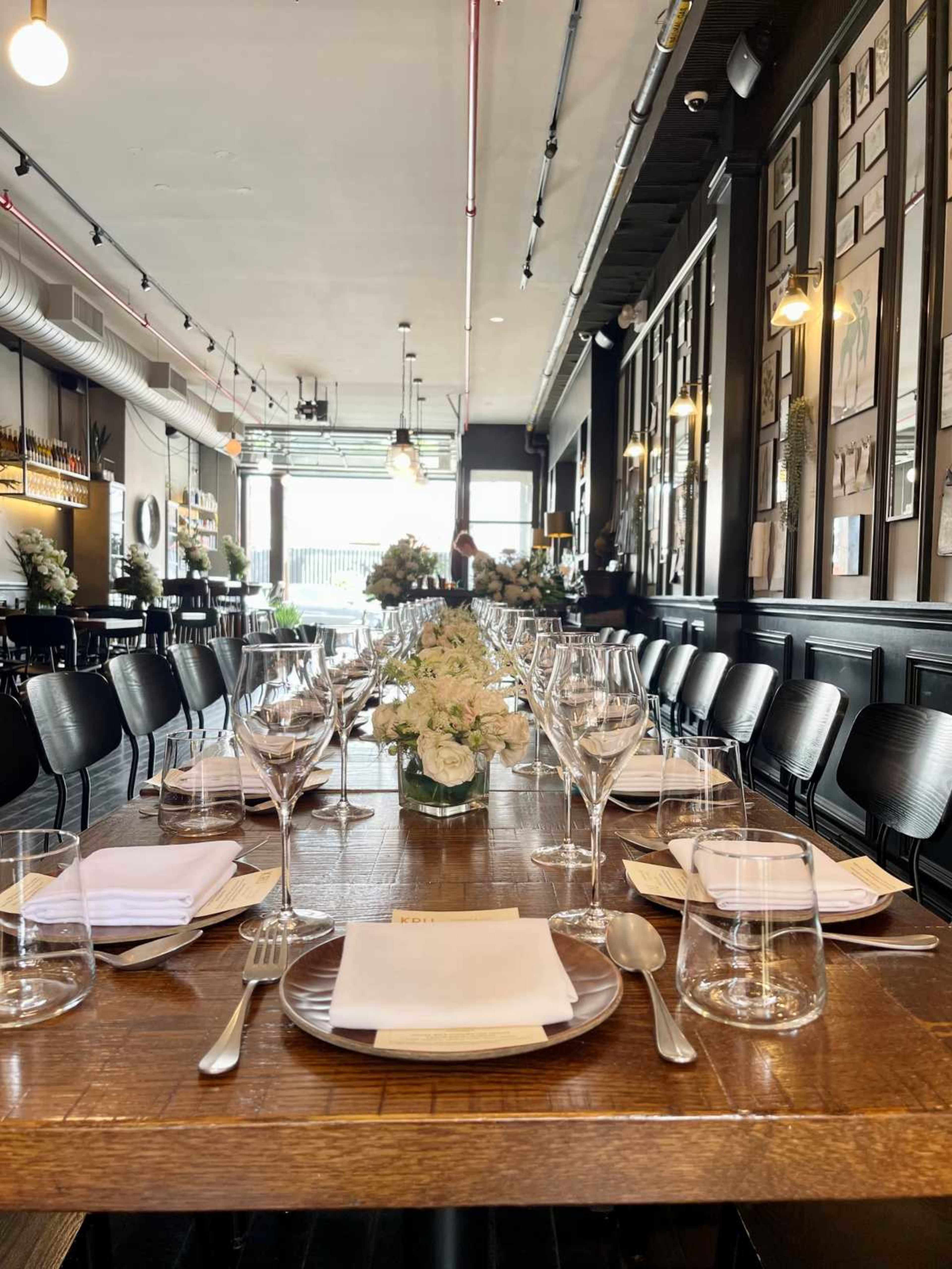 The image shows a long wooden dining table set with glassware and white napkins, adorned with a floral centerpiece, inside a modern restaurant.