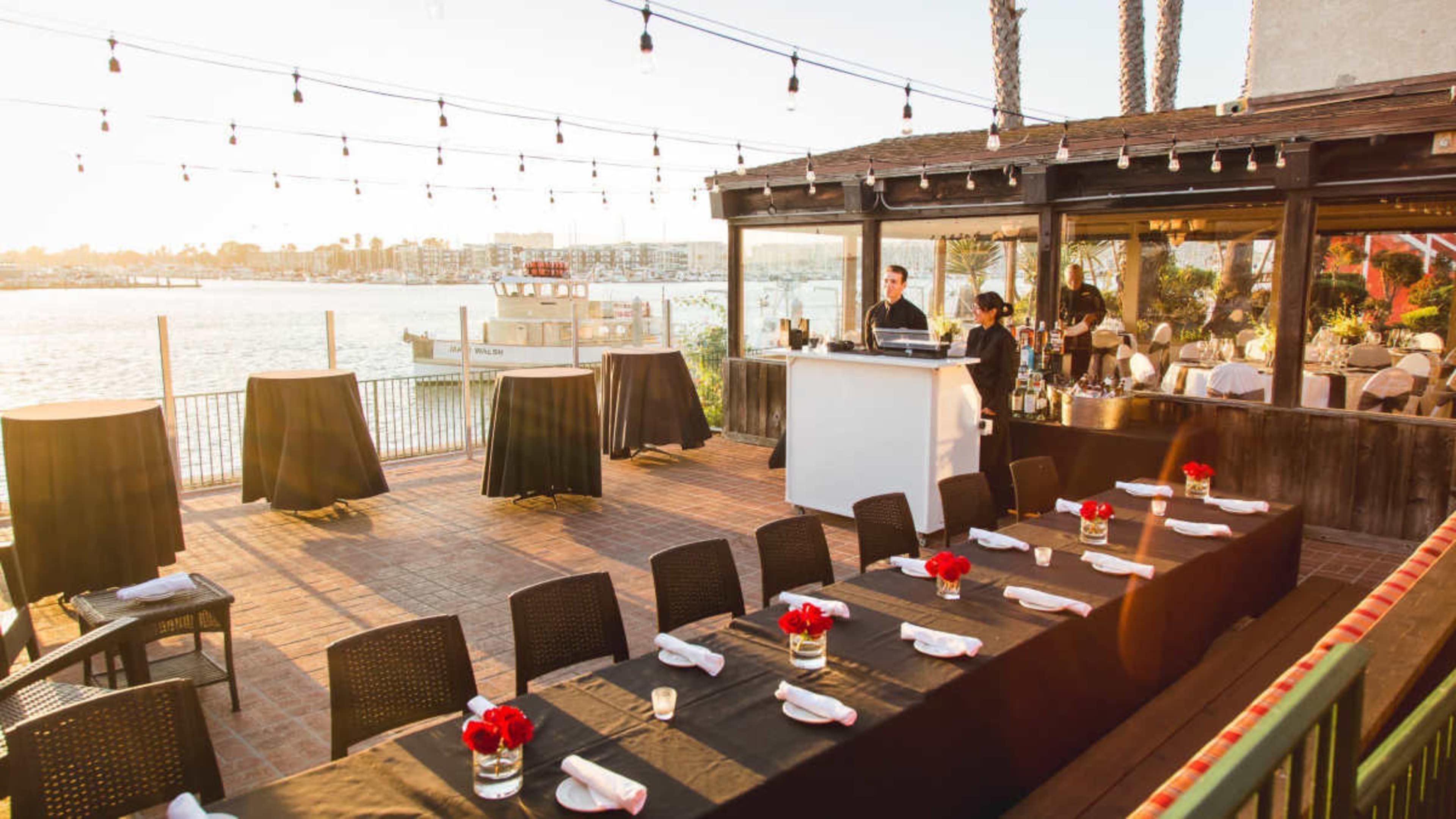 The image shows a rooftop restaurant terrace with tables set for dining, a bar area, and views of a waterfront at sunset.