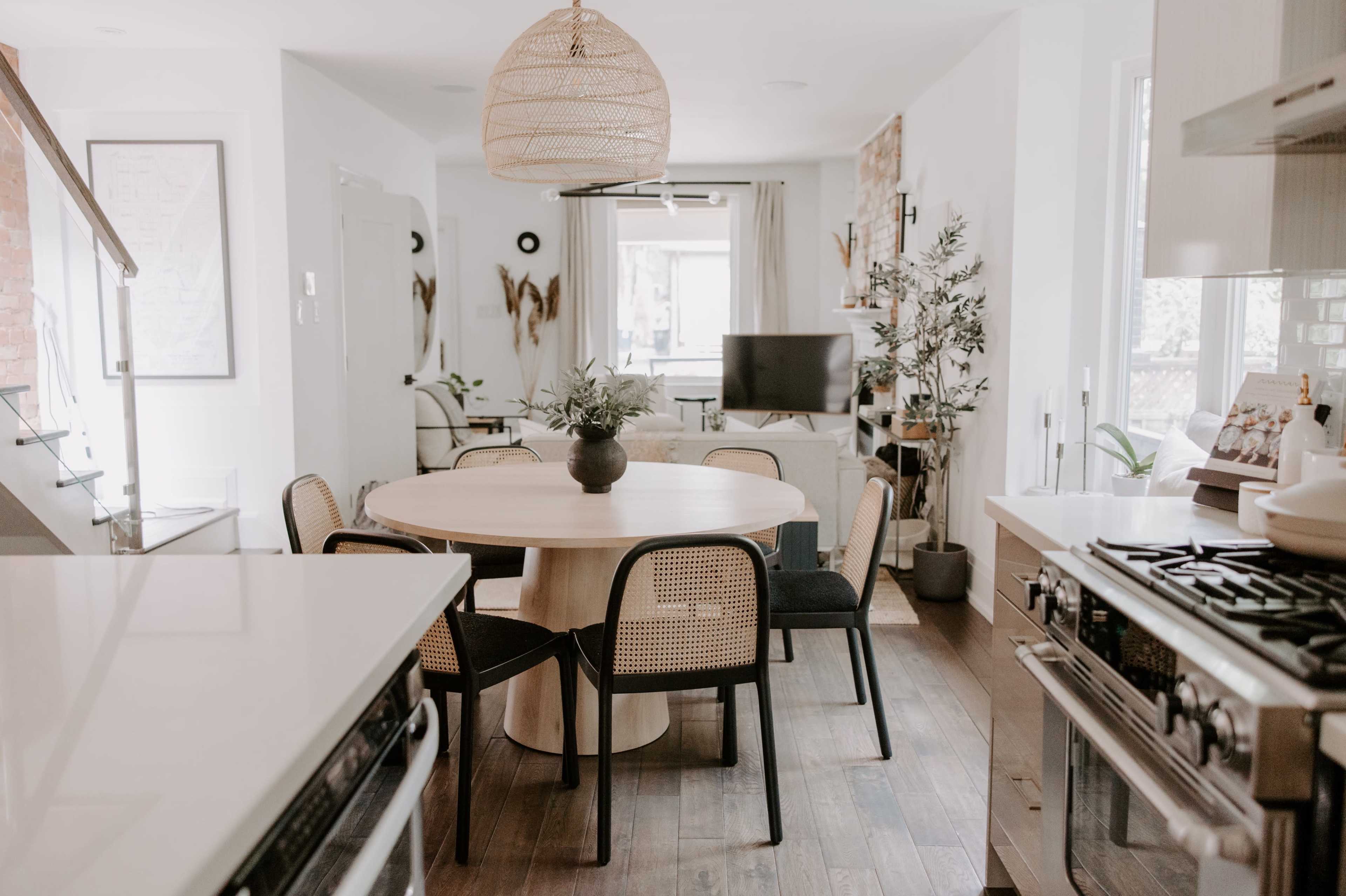 A modern kitchen and dining area features a round wooden table surrounded by black and woven chairs, with a large window and plants adding to the decor.