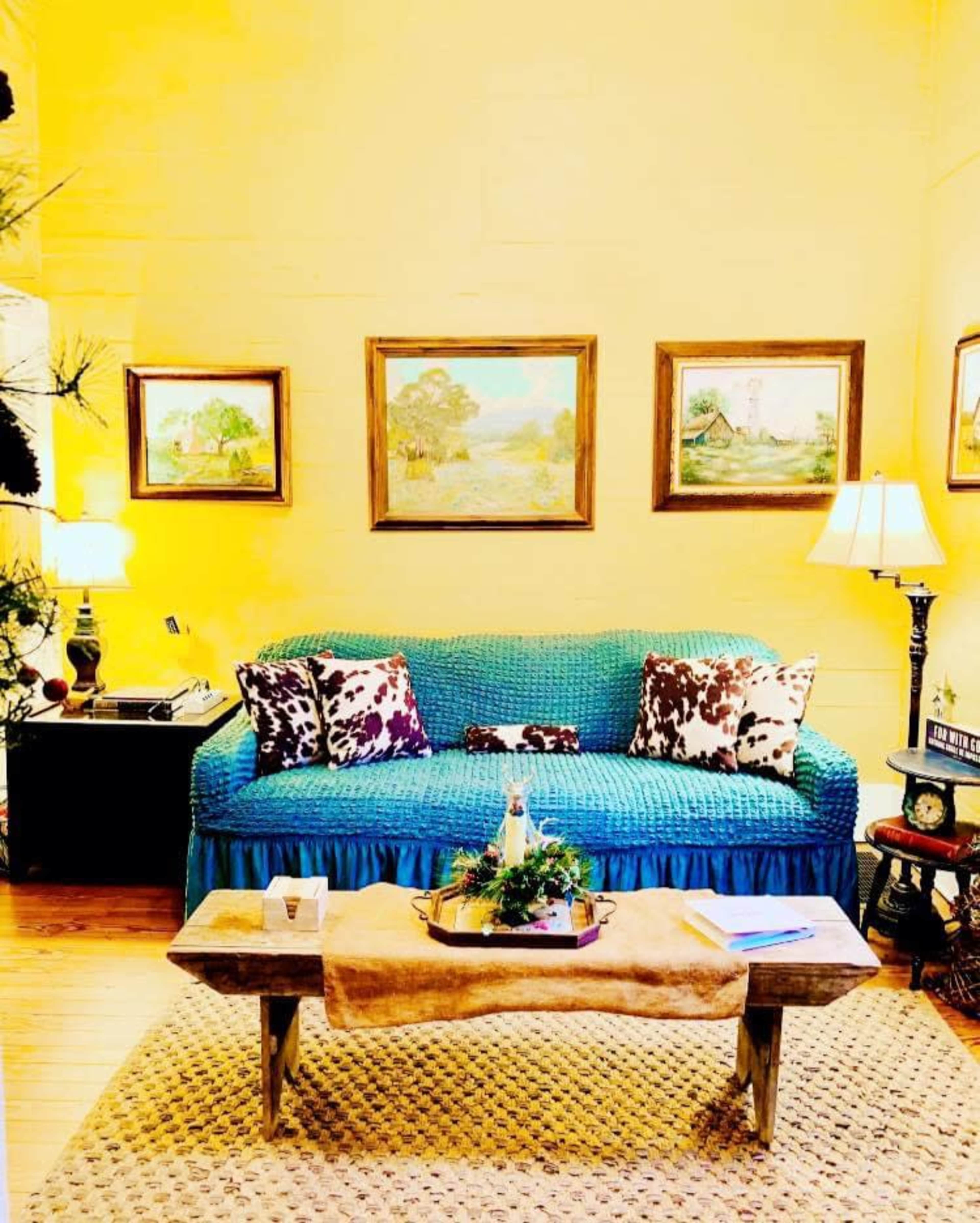 The scene shows a cozy living room with a bright yellow wall, an aqua-colored couch adorned with patterned pillows, two lamps, and artwork hanging above, all complemented by a rustic wooden coffee table and a woven rug.