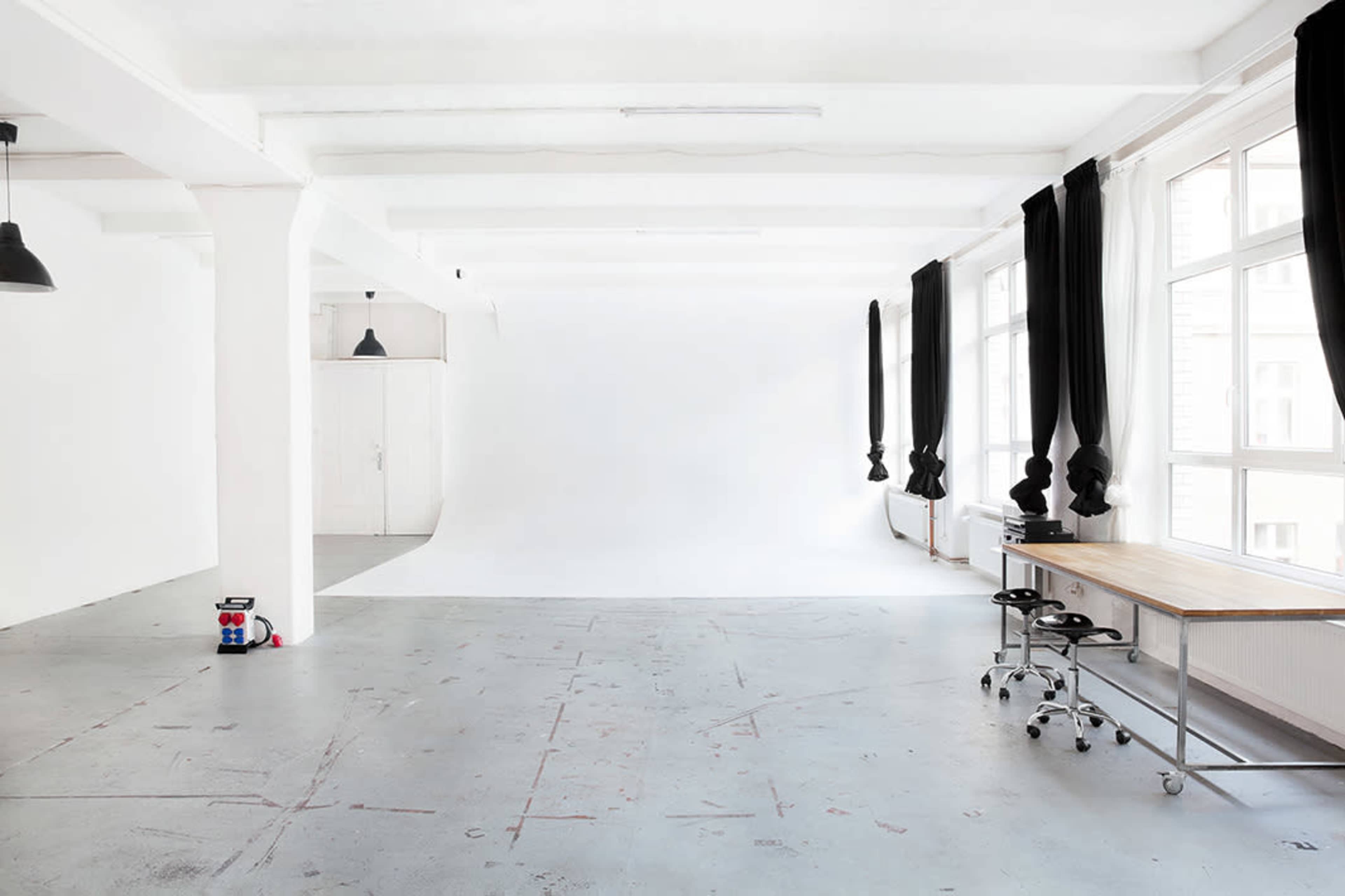 A spacious, empty studio with a white backdrop, large windows, and a wooden desk on wheels.