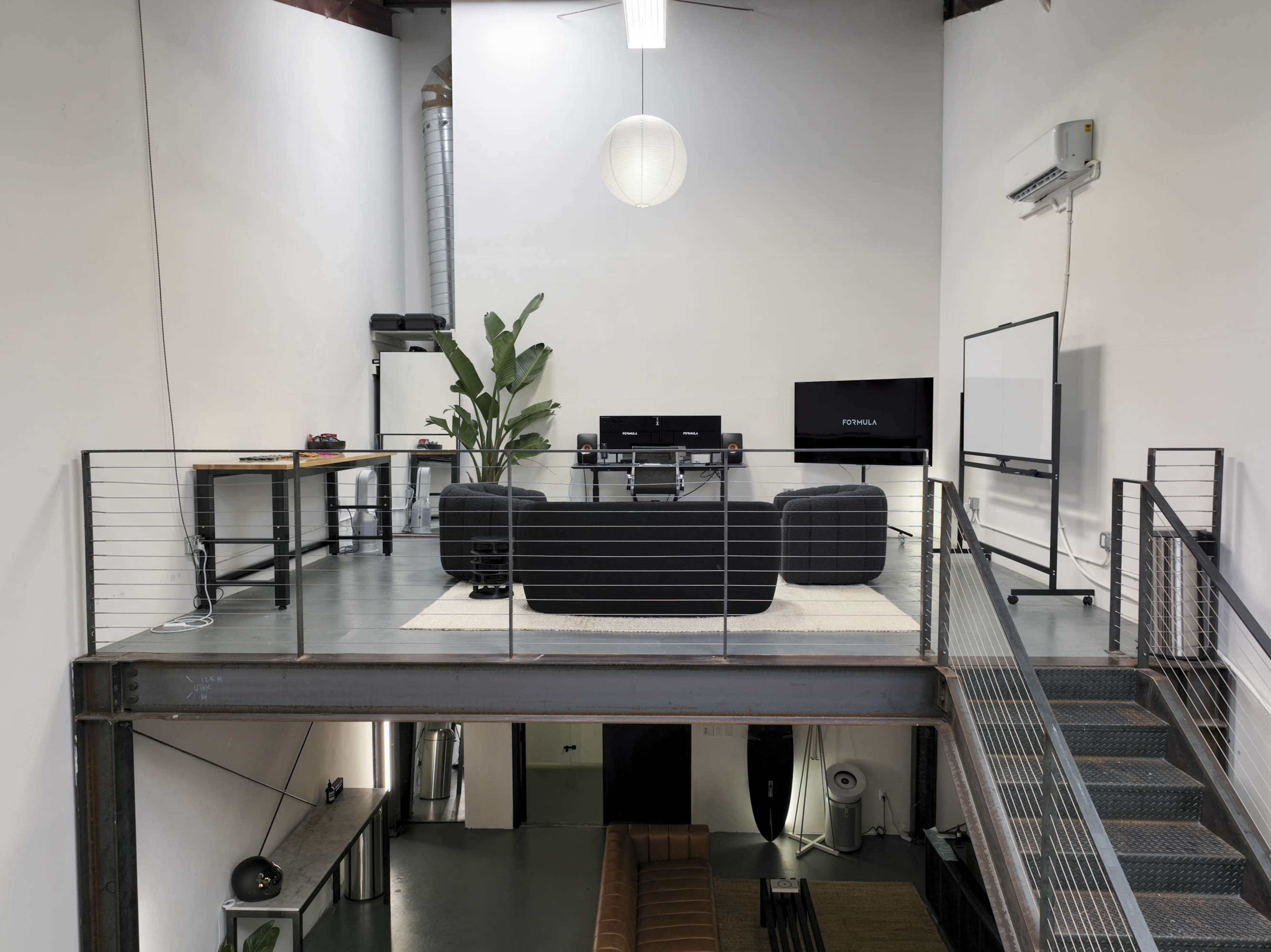 A contemporary open loft space with a second-floor seating area, featuring a black couch, computer setup, and a potted plant.