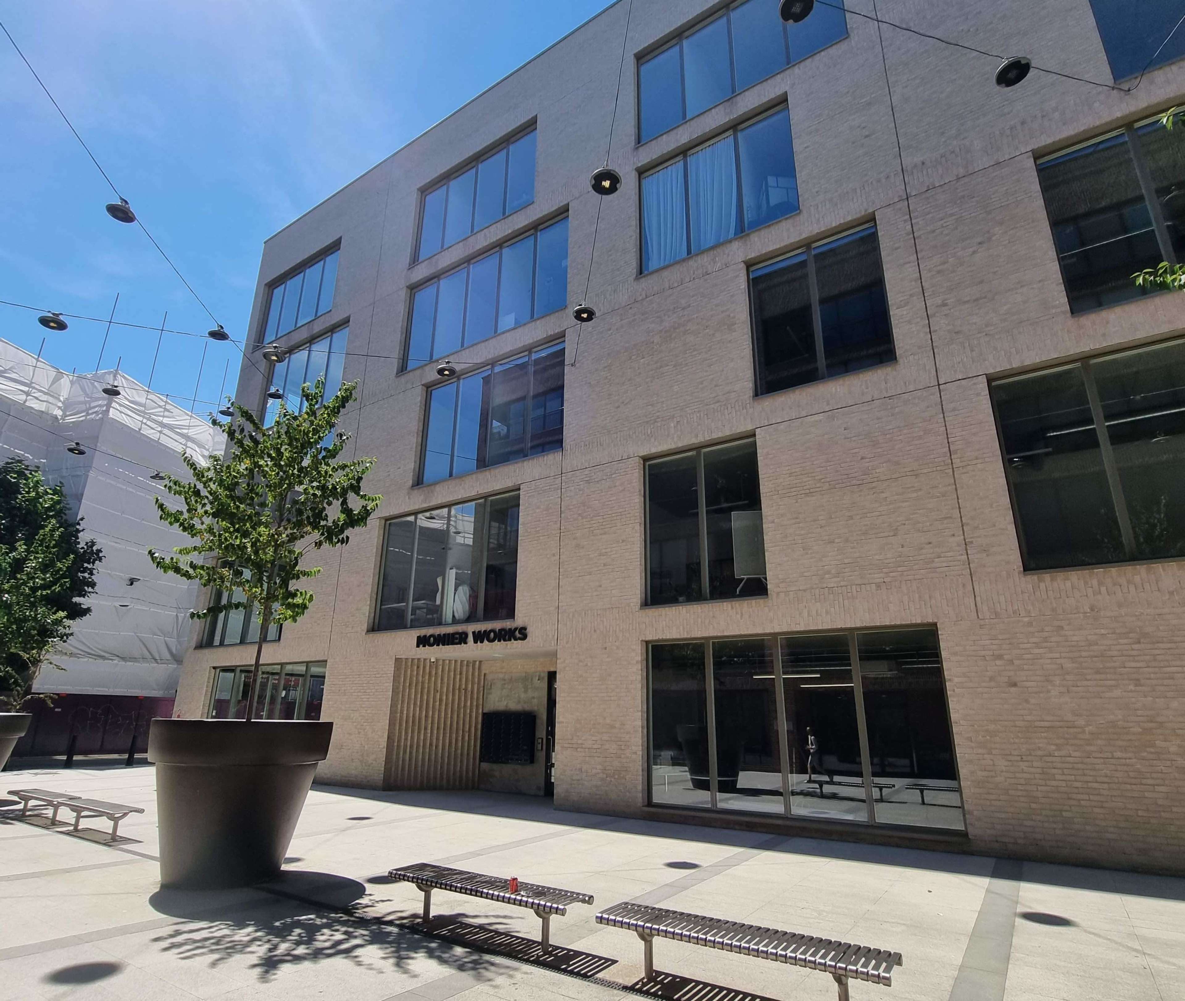 A modern building with large windows features a courtyard that includes a tree and several benches.
