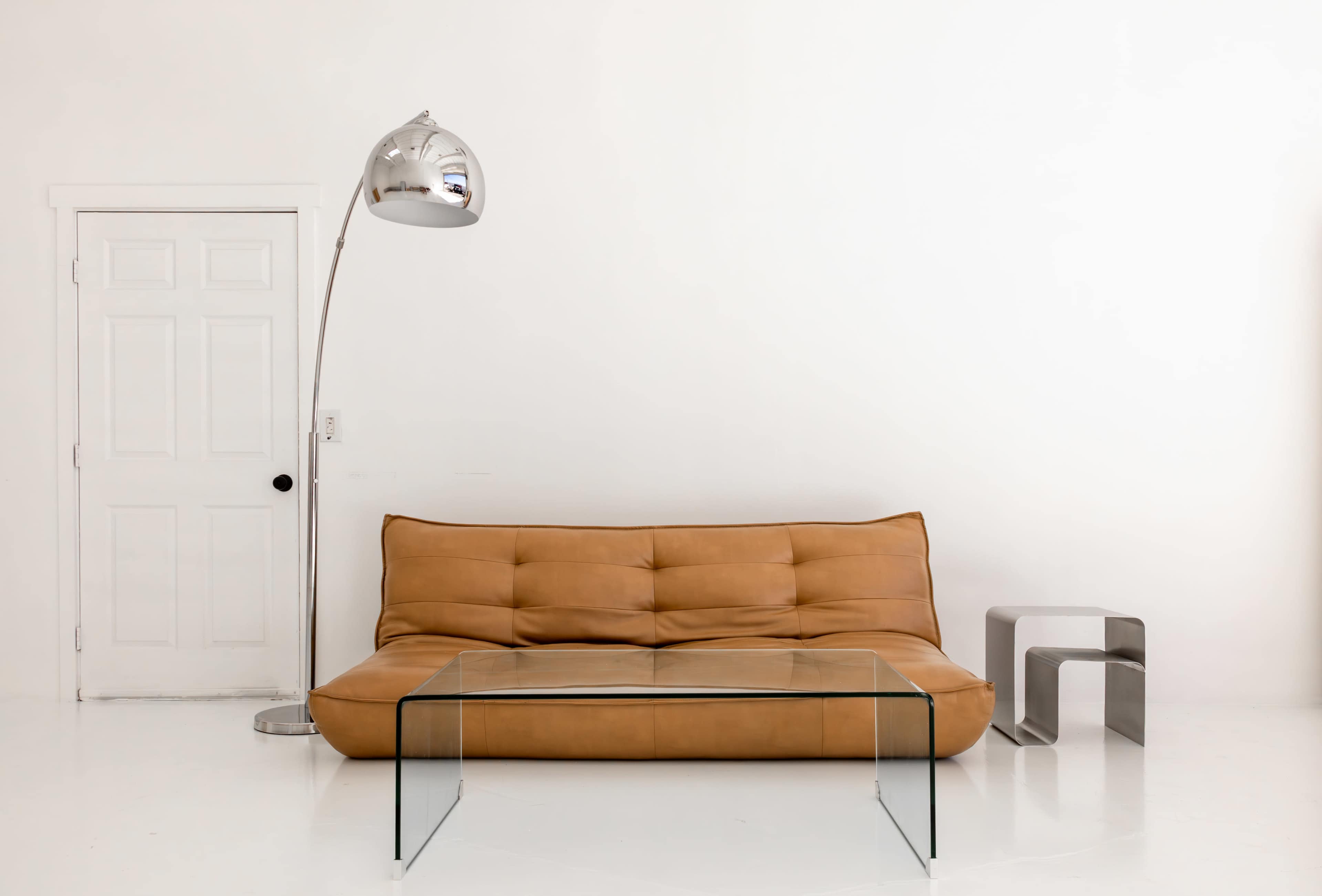 The image shows a modern living room setup featuring a tan sofa, a clear glass coffee table, and a chrome floor lamp against a white wall.