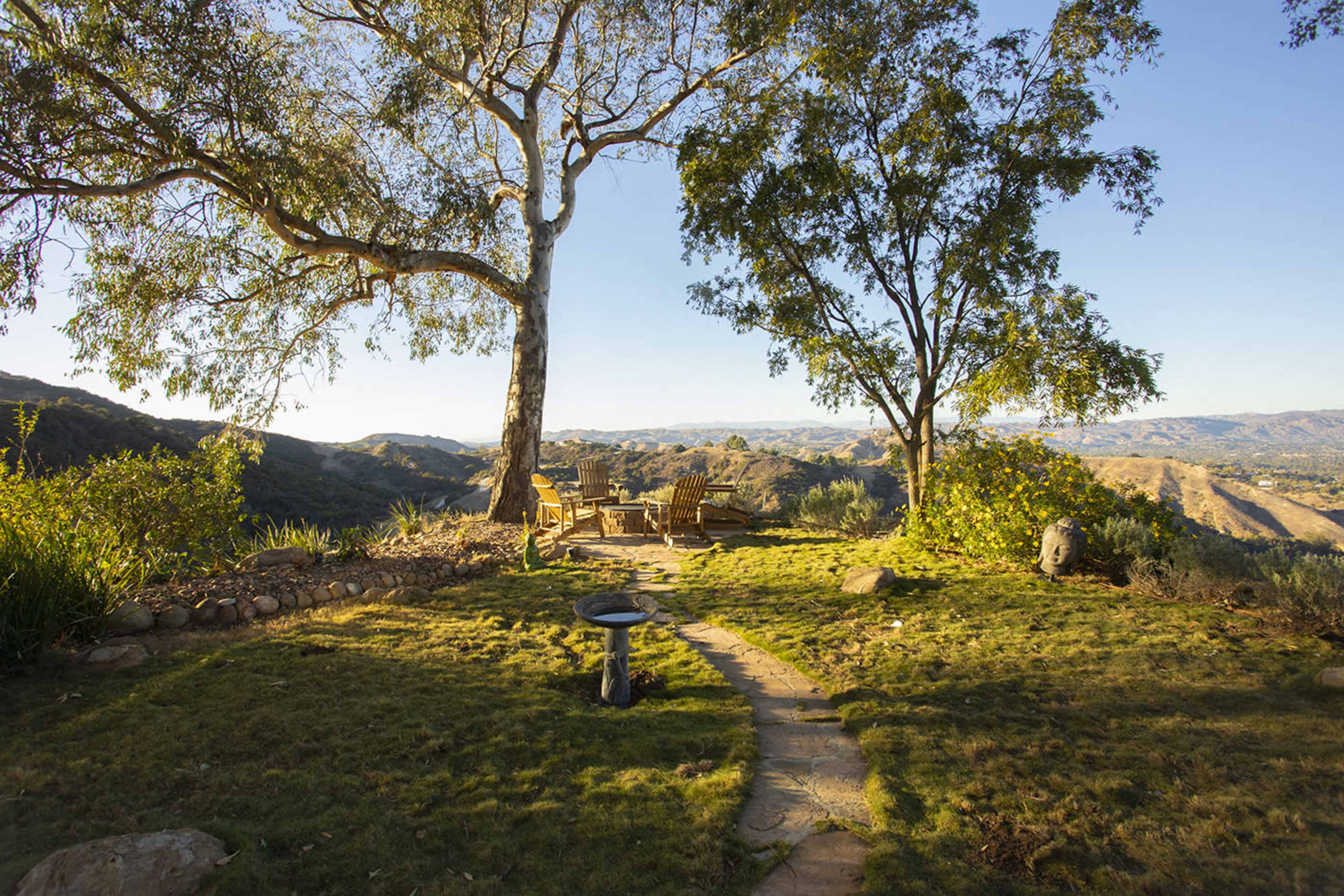 The image shows a serene outdoor setting with two trees, a stone pathway, and a wooden seating area overlooking rolling hills.