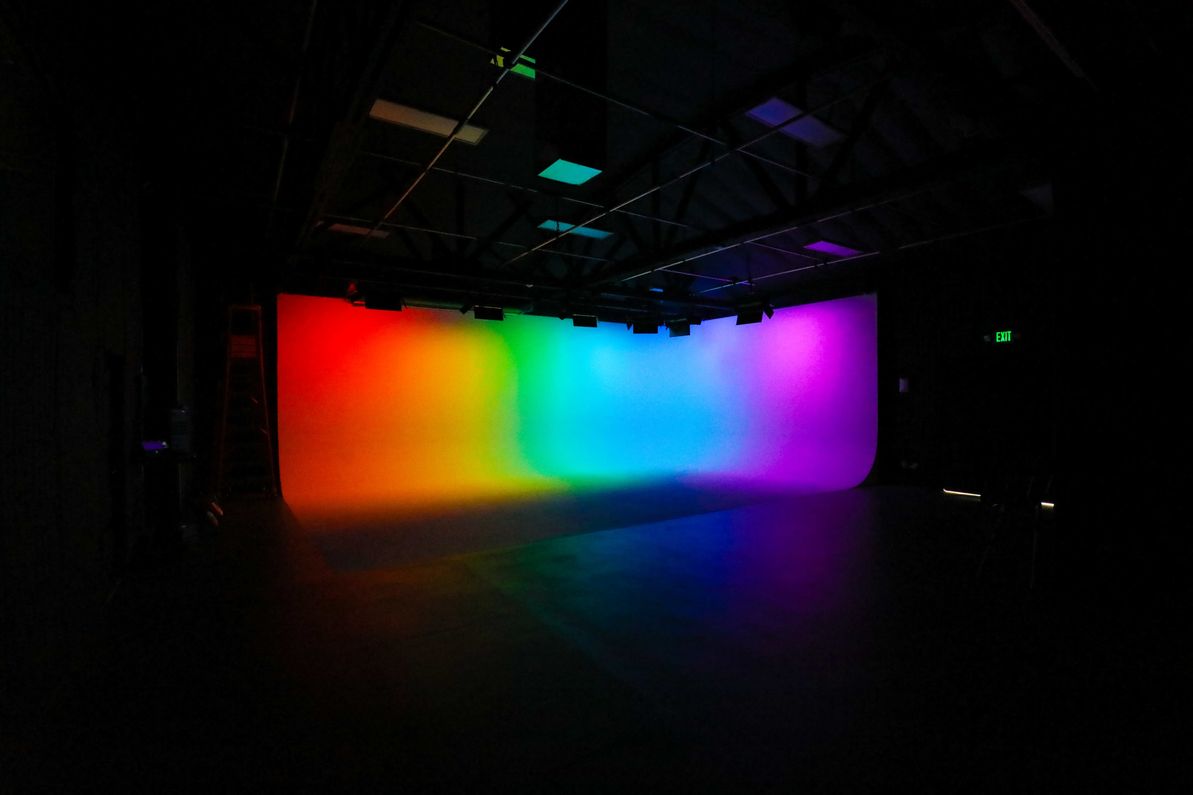 A dimly lit studio space with a backdrop that transitions smoothly from red to orange, yellow, green, blue, and purple.