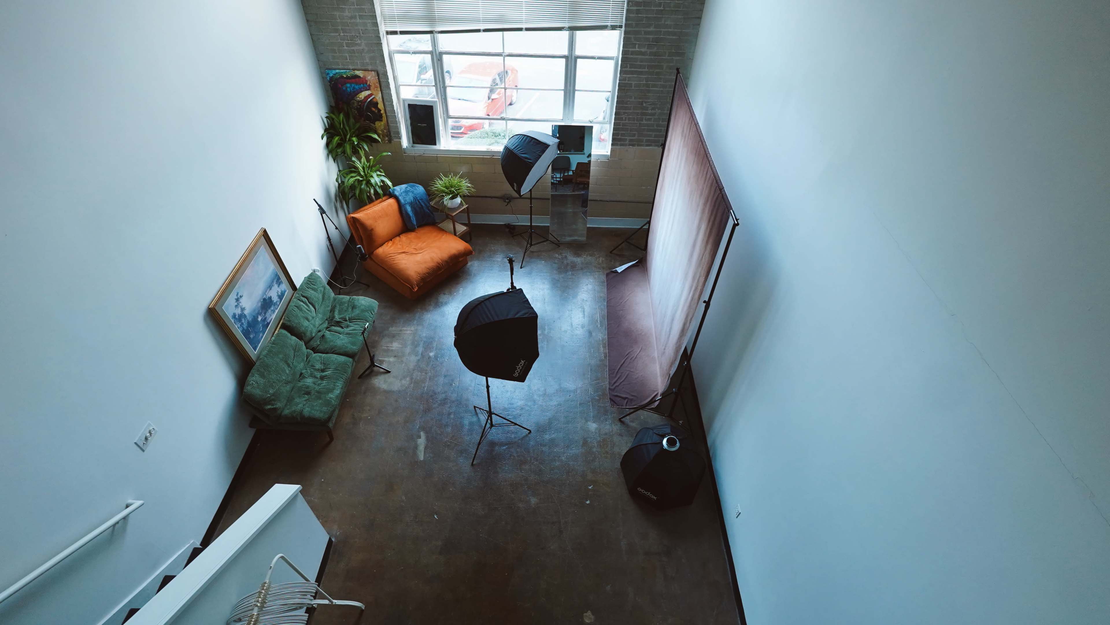 The image shows a spacious room with a large window, a green couch, an orange chair, and several photography lights set up.