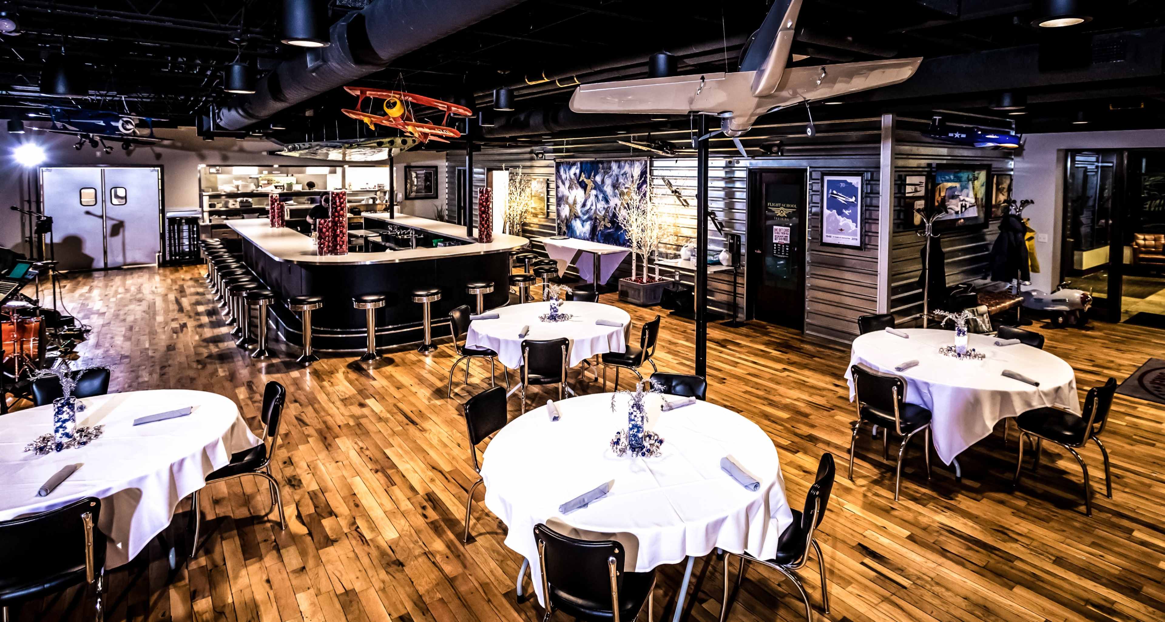 A modern restaurant interior with circular tables covered in white tablecloths, a bar area, and aviation-themed decor including a model airplane hanging from the ceiling.
