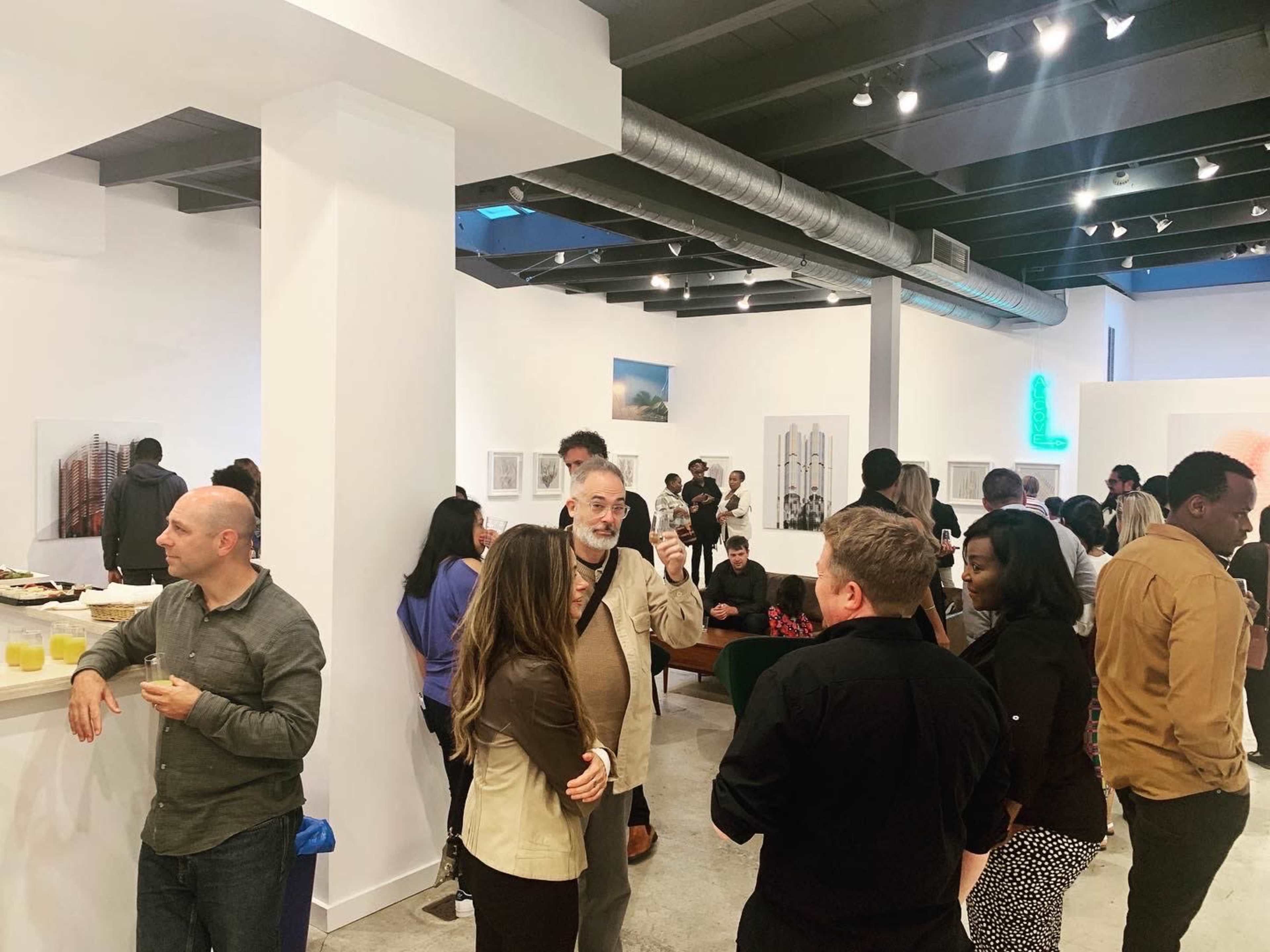 A diverse group of people engages in conversation at an art gallery opening with artworks displayed on the walls.