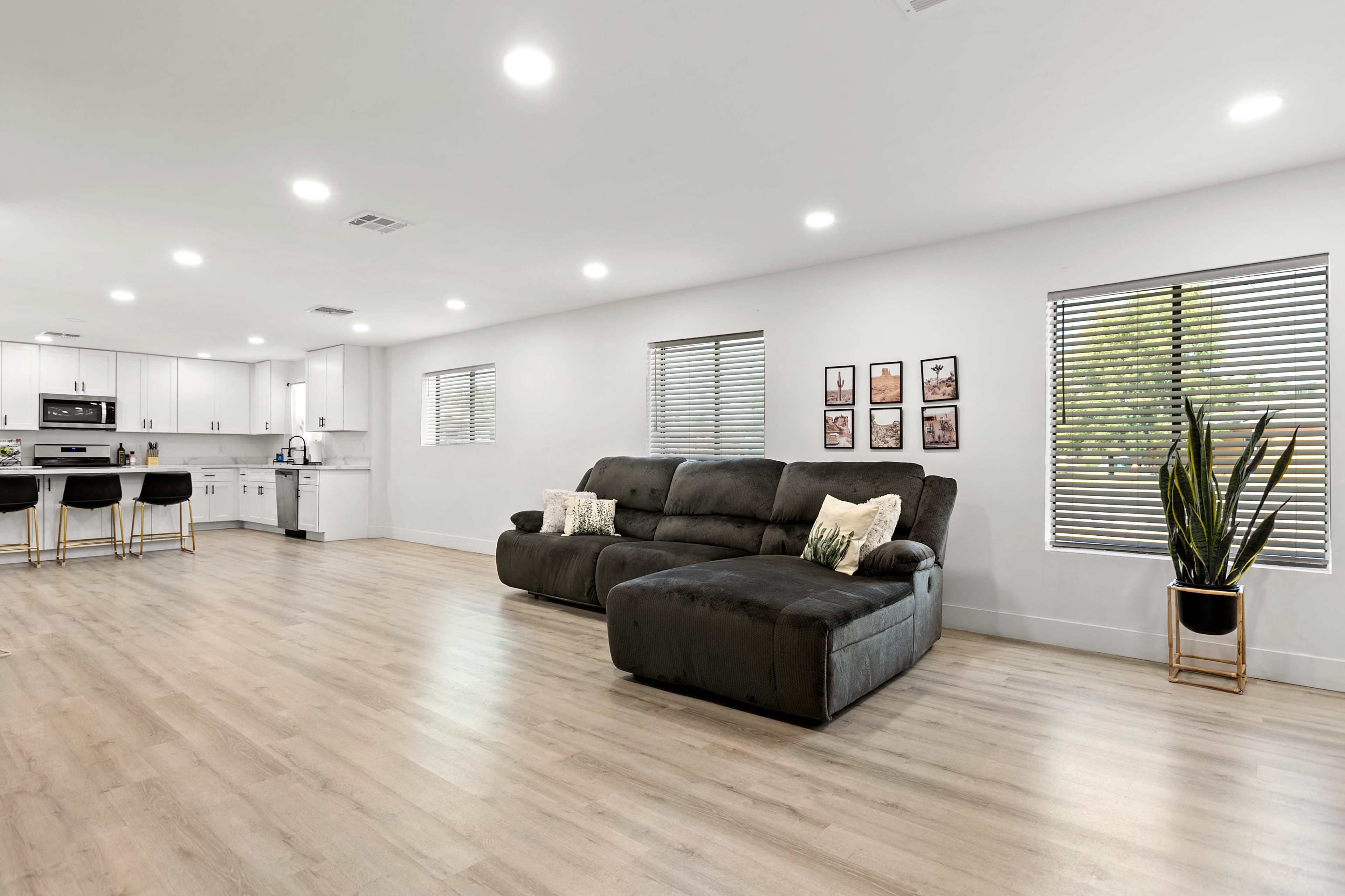 The image shows a modern living space featuring a dark sectional sofa, light wood flooring, and large windows with blinds.