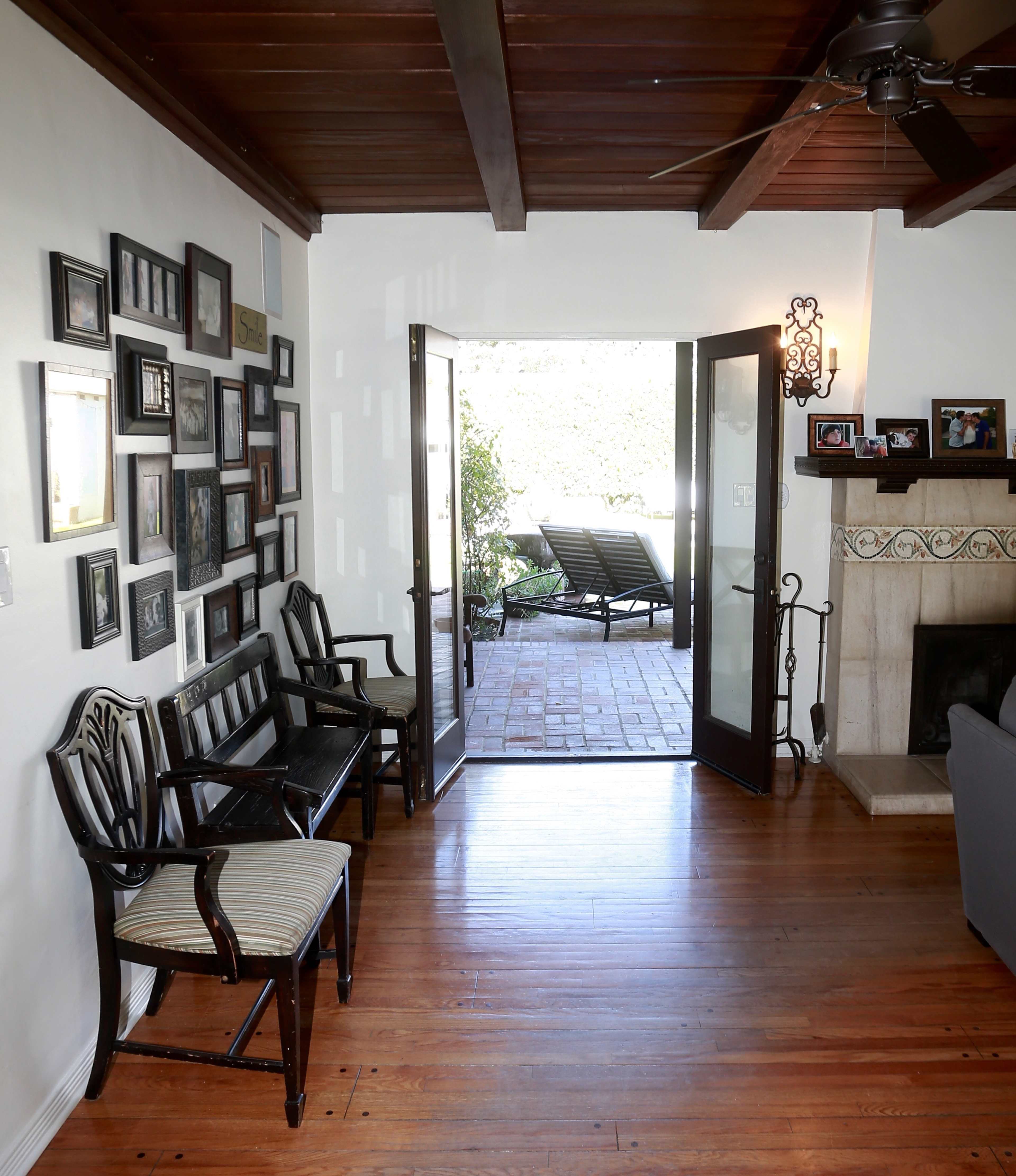 The image shows a living room with wooden floors, a beamed ceiling, and a doorway leading to an outdoor patio with lounge chairs.