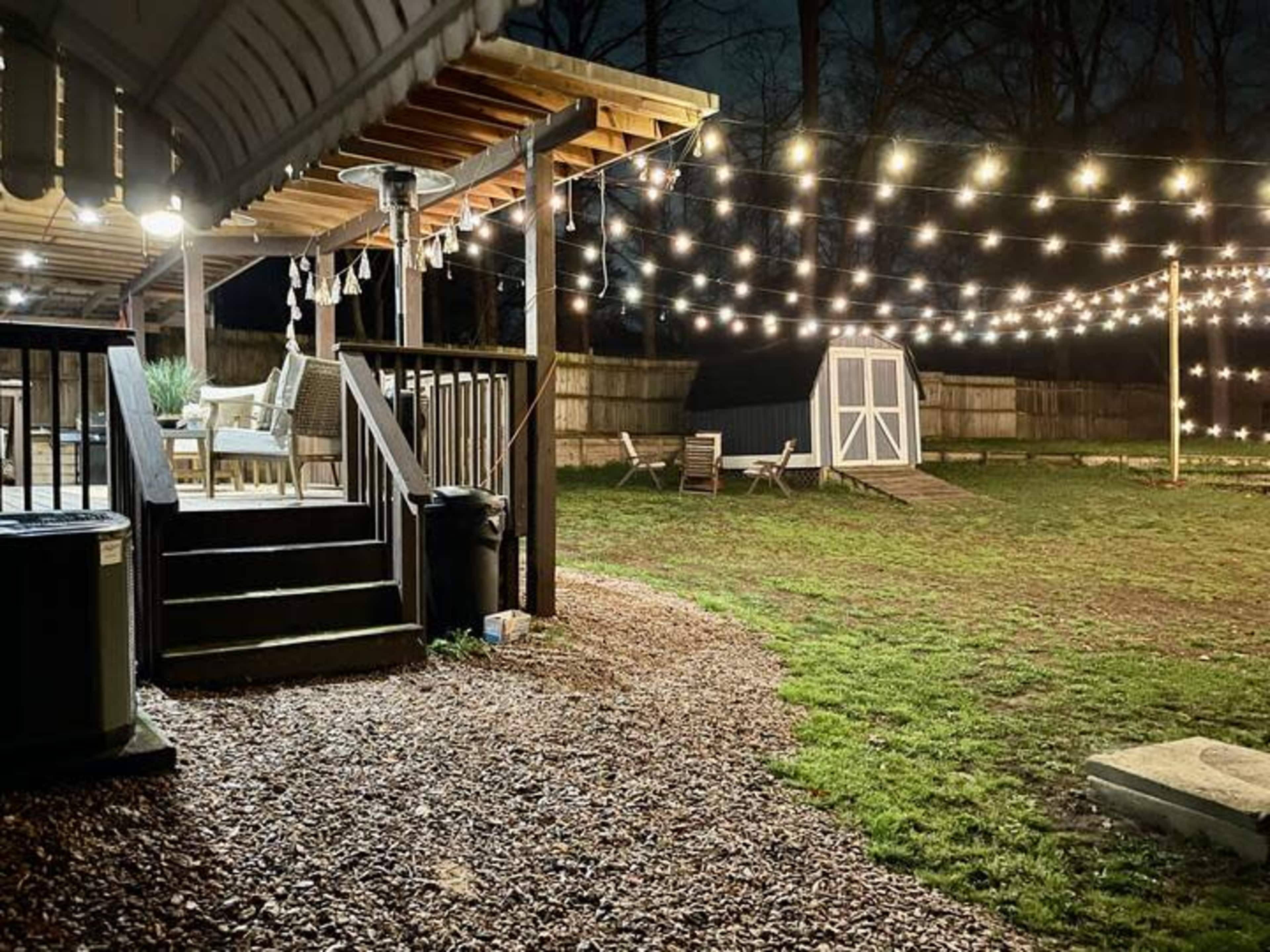 A backyard at night, featuring a patio with string lights overhead and a shed in the distance.