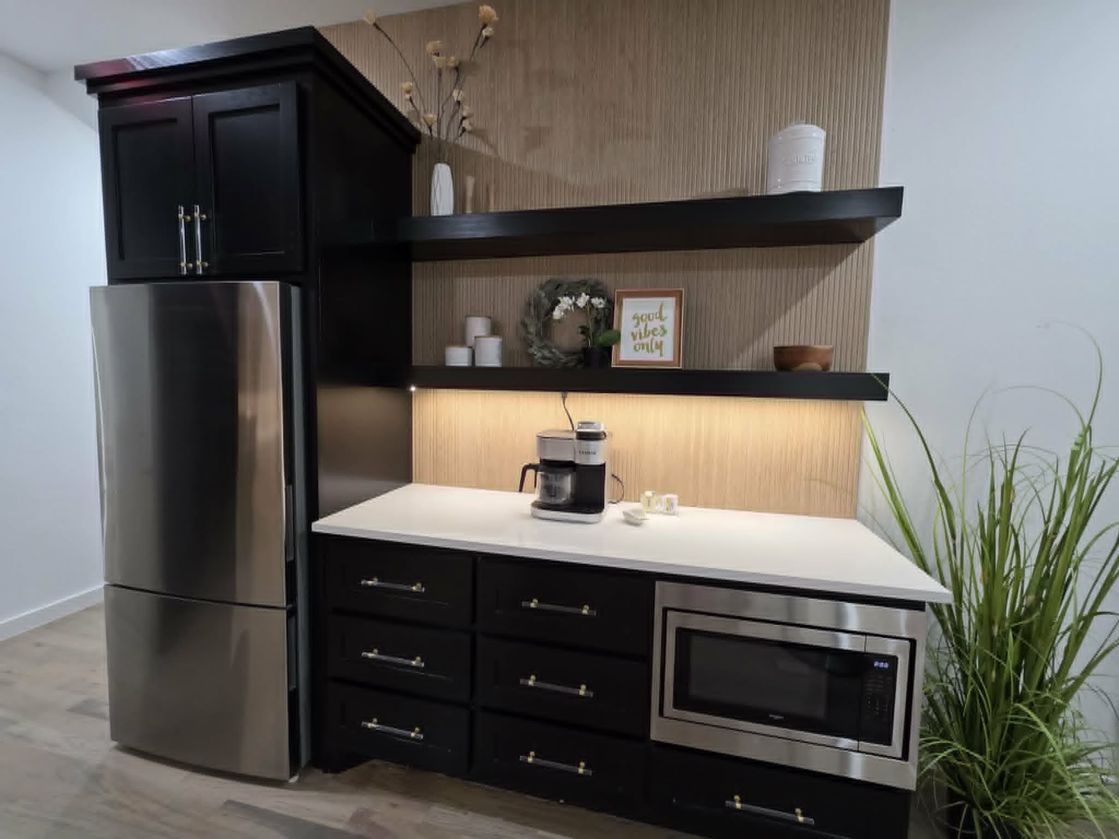 The image shows a modern kitchen featuring a stainless steel refrigerator, black cabinetry, a countertop with a coffee maker, and decorative shelves.
