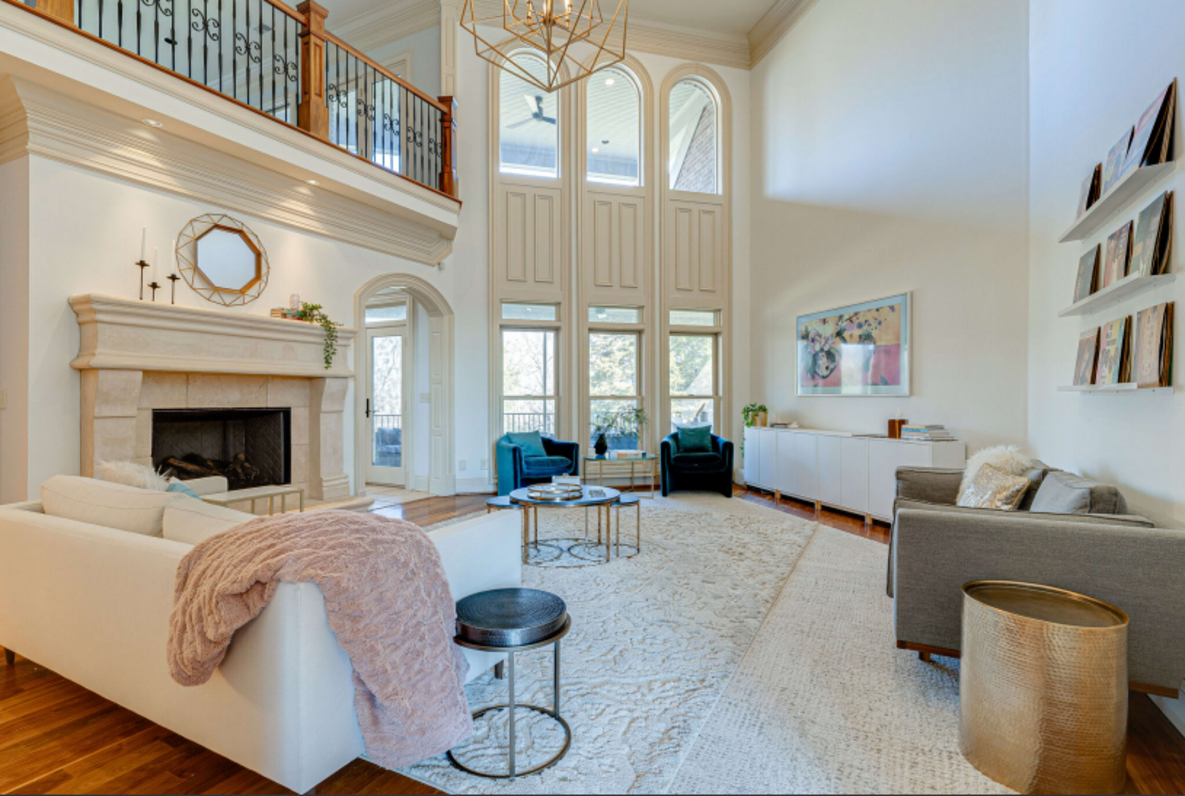 The image shows a spacious living room with high ceilings, a stone fireplace, large windows, and modern furniture arranged around a central area rug.