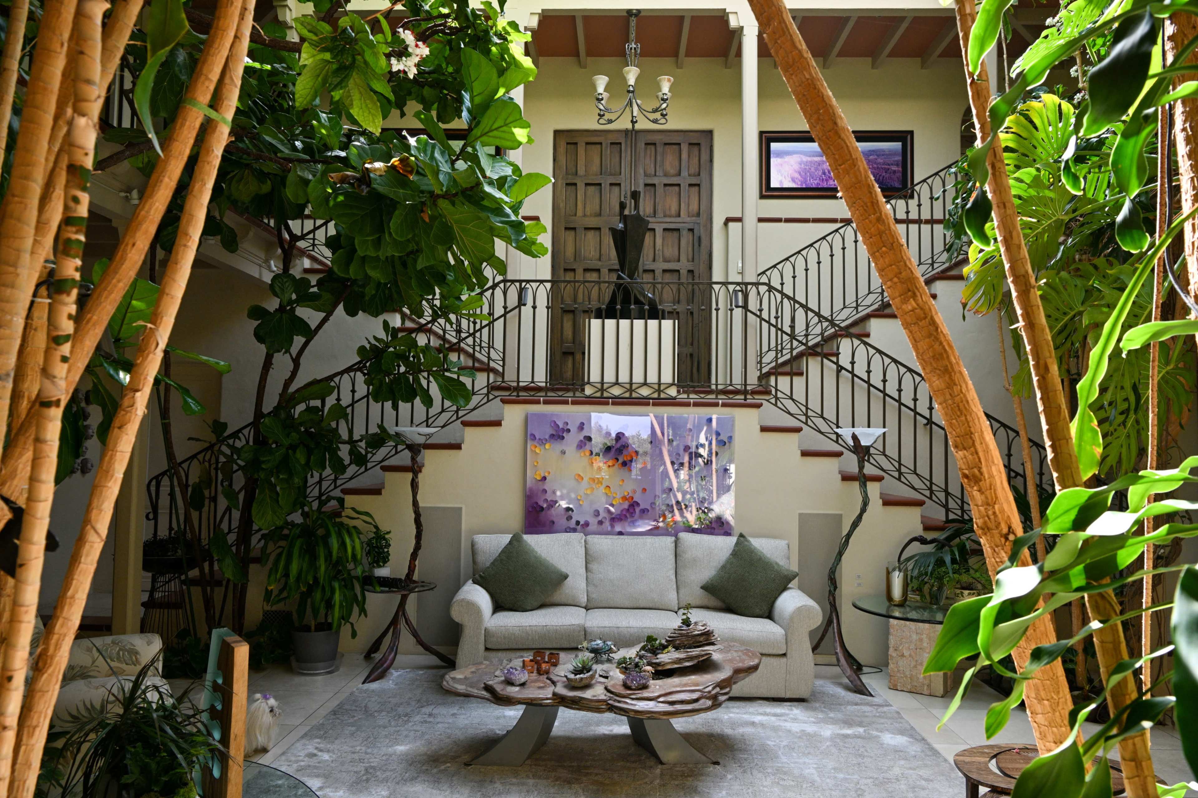 The image shows a cozy indoor space featuring a sofa and a unique coffee table, surrounded by lush greenery and a staircase leading to an upper floor.