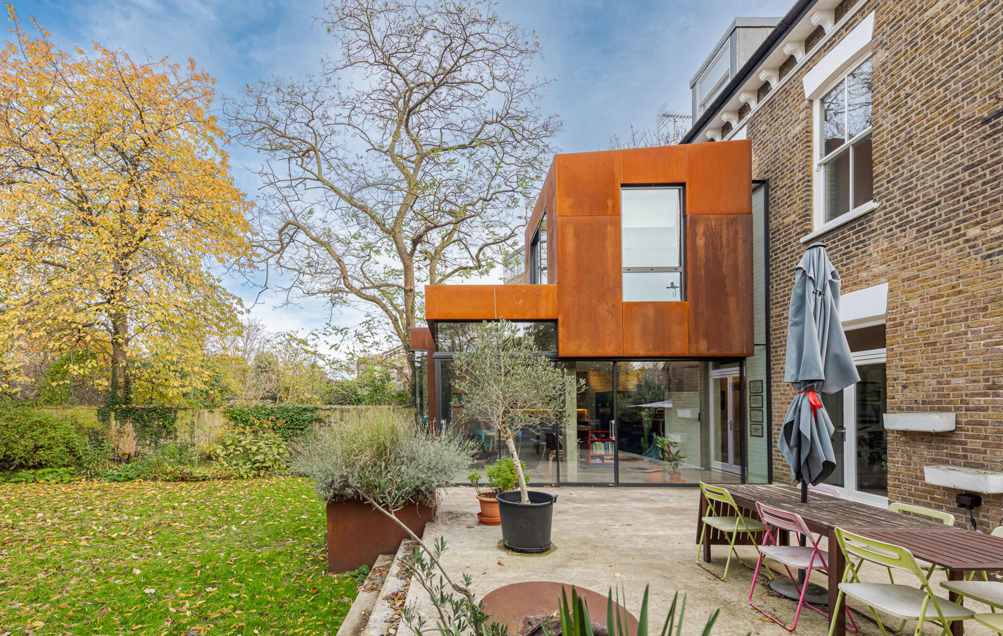 The image shows a modern house with a rusted metal extension and a patio surrounded by greenery and autumn leaves.