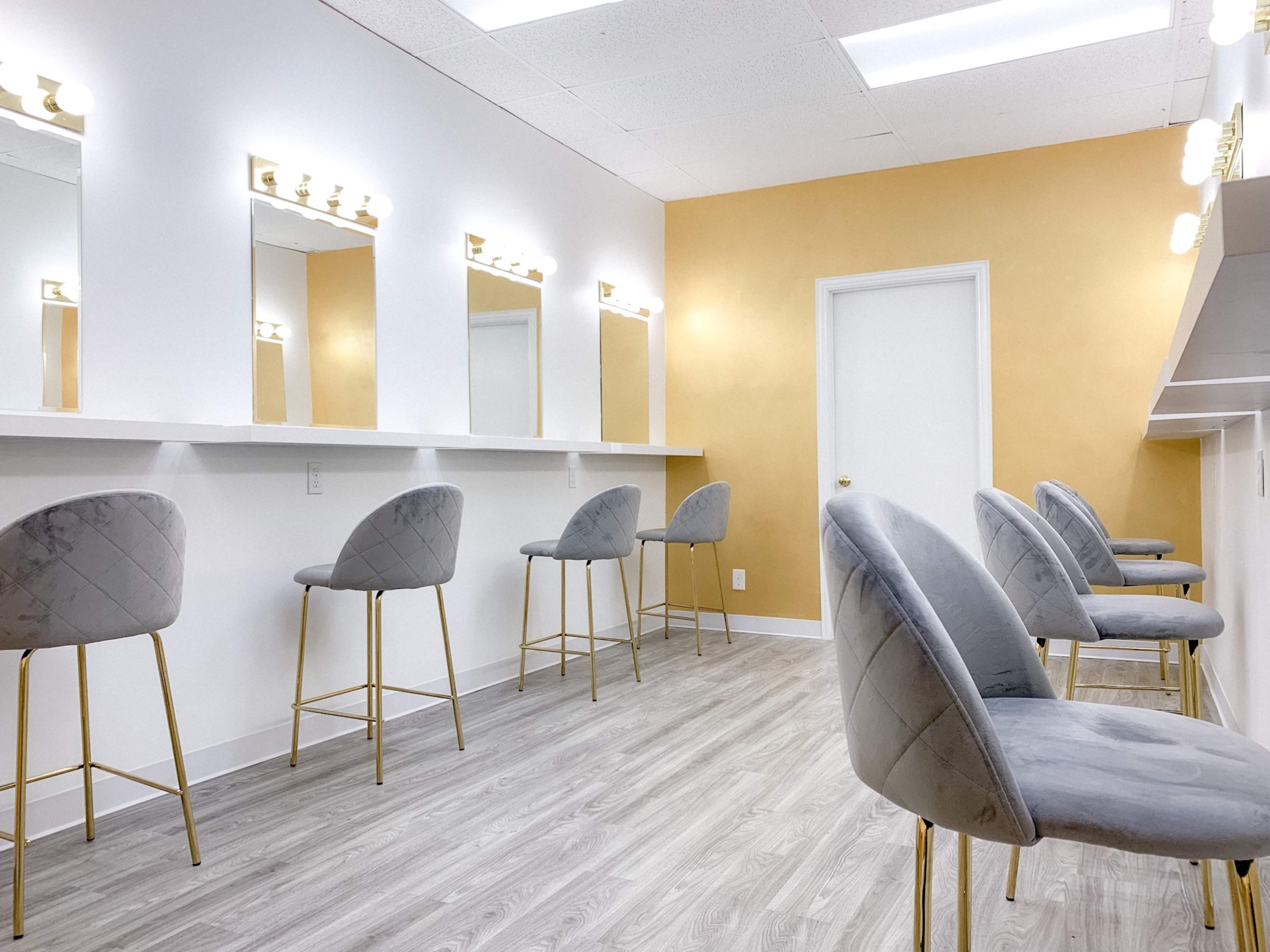A makeup station features several gray bar stools with gold legs arranged in front of illuminated mirrors against a gold-accented wall.