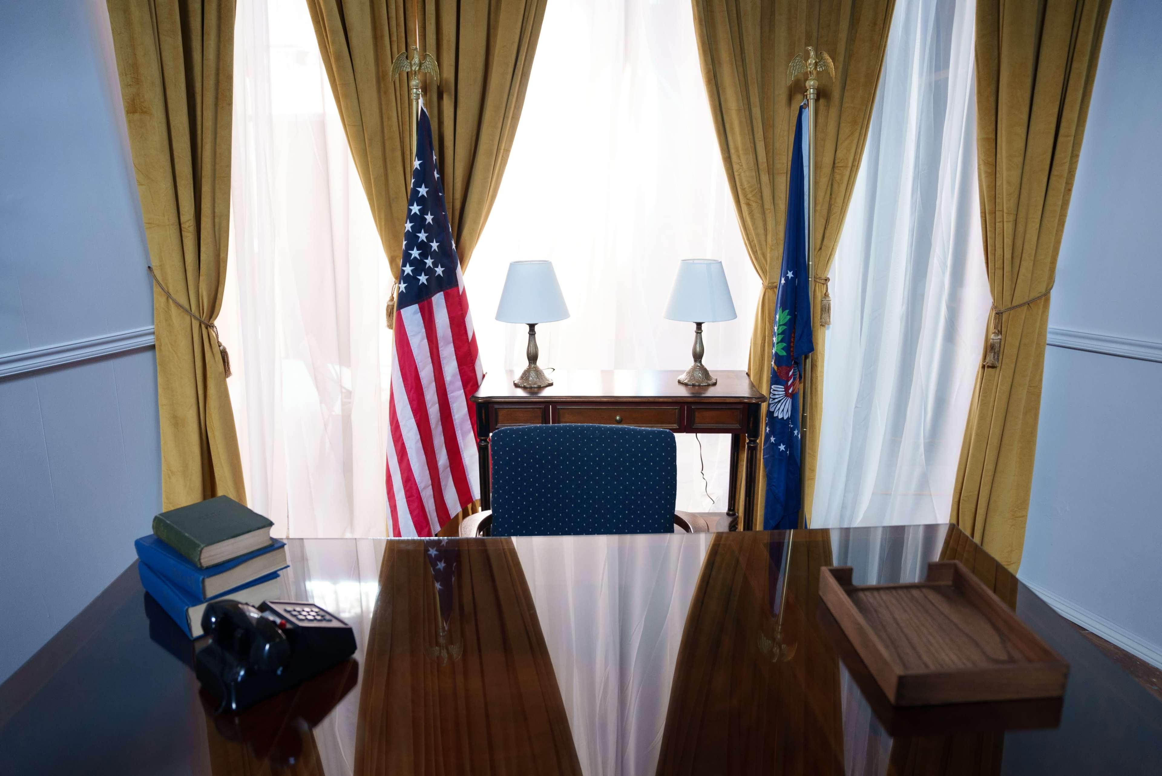 A presidential office is shown with an American flag, a state flag, a desk with a chair, two lamps, and a telephone.
