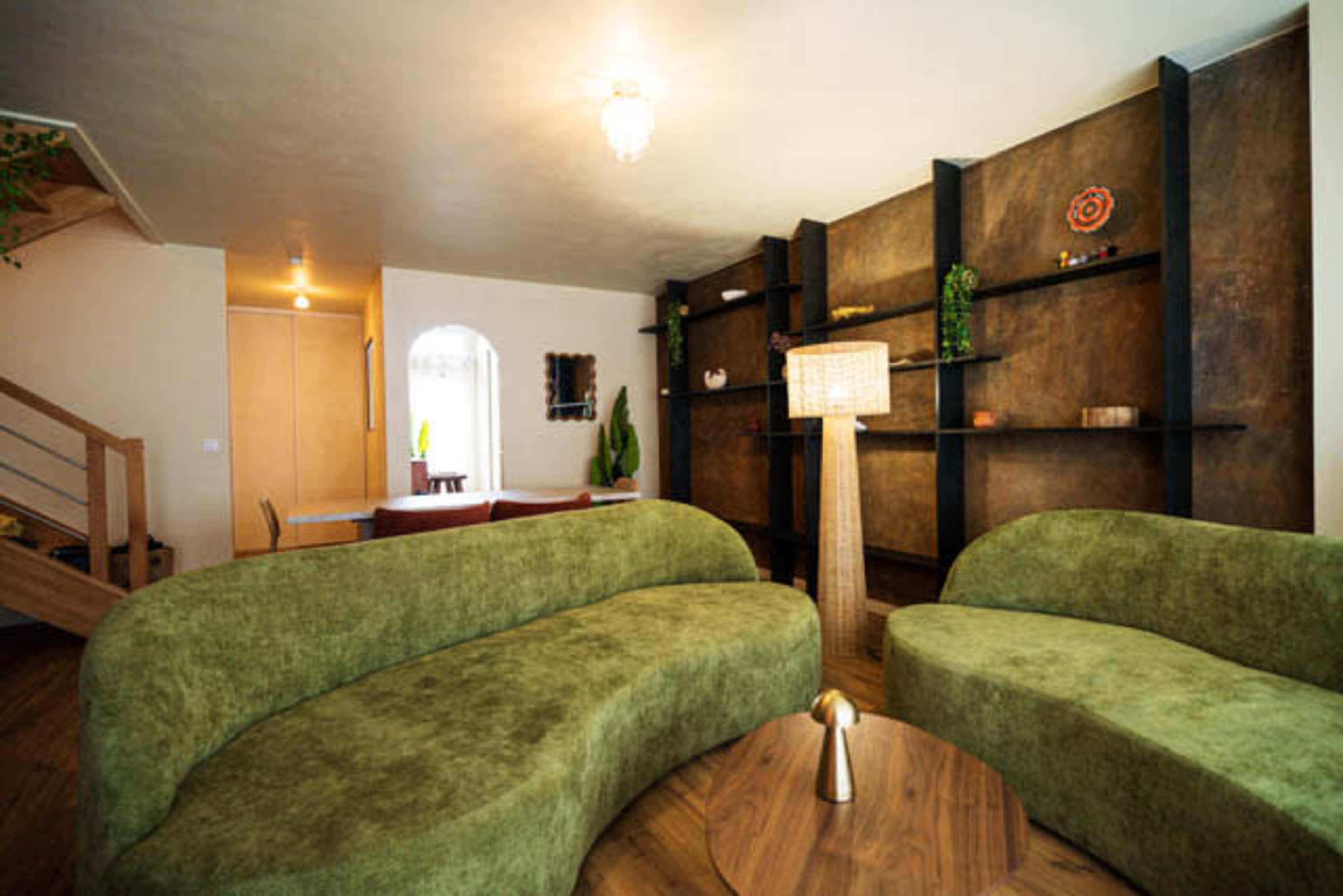 The image shows a modern living room with two curved green sofas facing a round wooden coffee table, and a textured brown wall with shelves and decorative items.